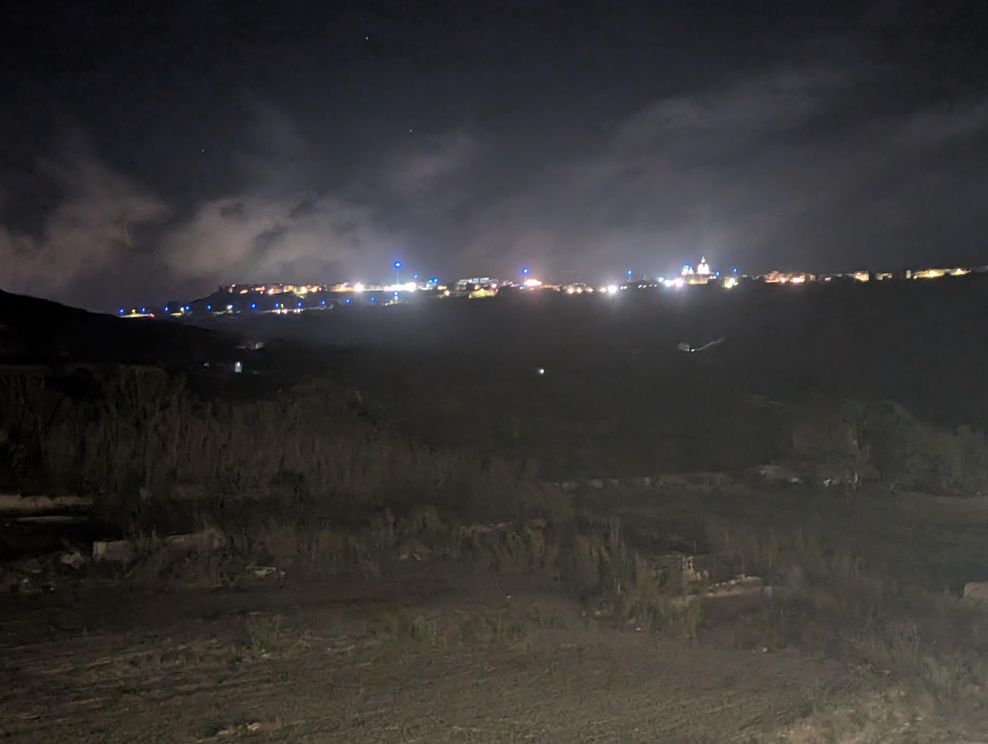 Photo of a village in the night
Dark hills before and illuminated village and sky.
A line of blue light is visible above the vilage