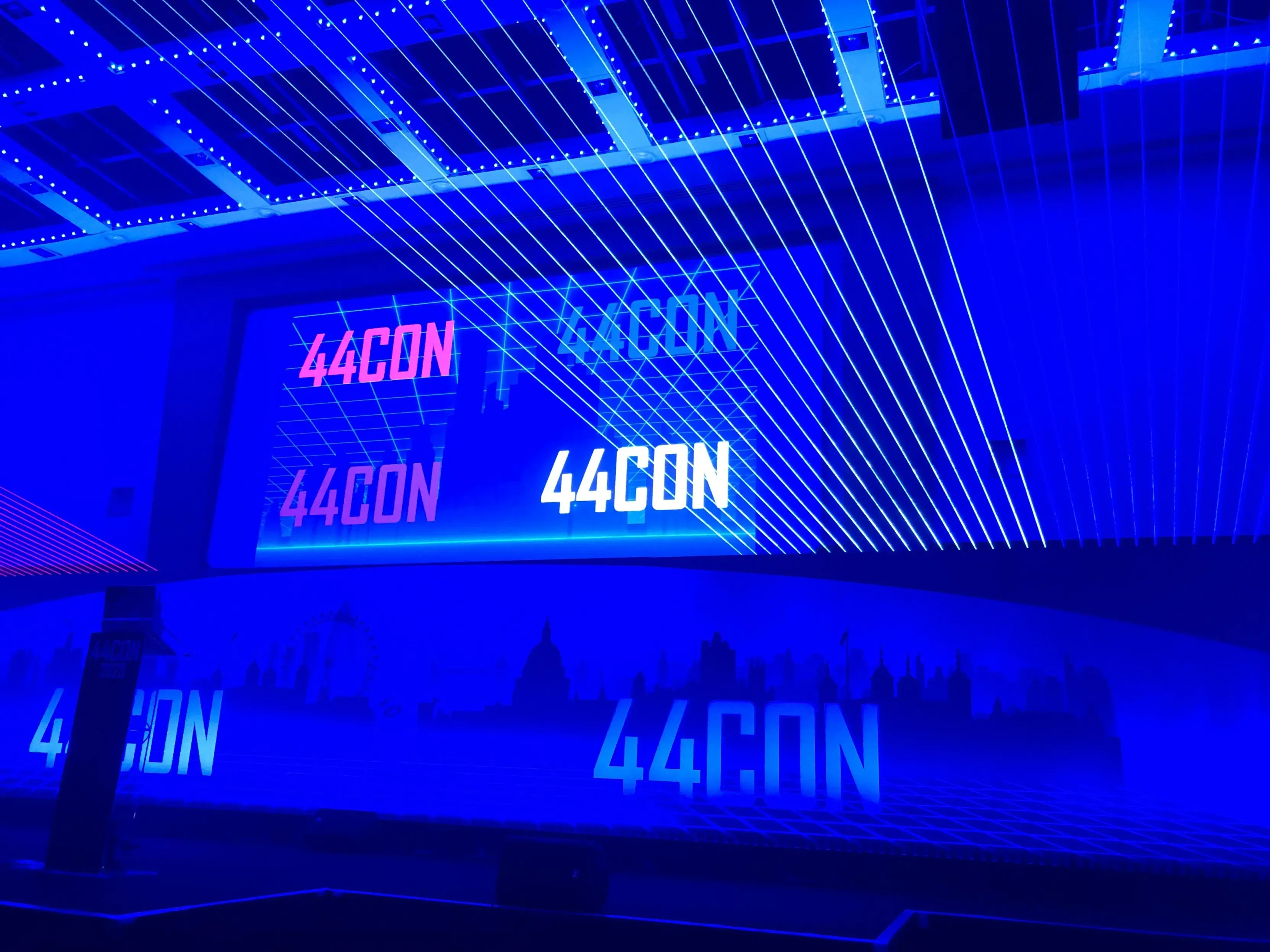 A dark conference hall lit in deep blue, with green laser lines fanning across the ceiling toward a large screen displaying the '44CON' logo repeated in pink, purple, and white. A London skyline silhouette and additional '44CON' branding appear along the stage below.
