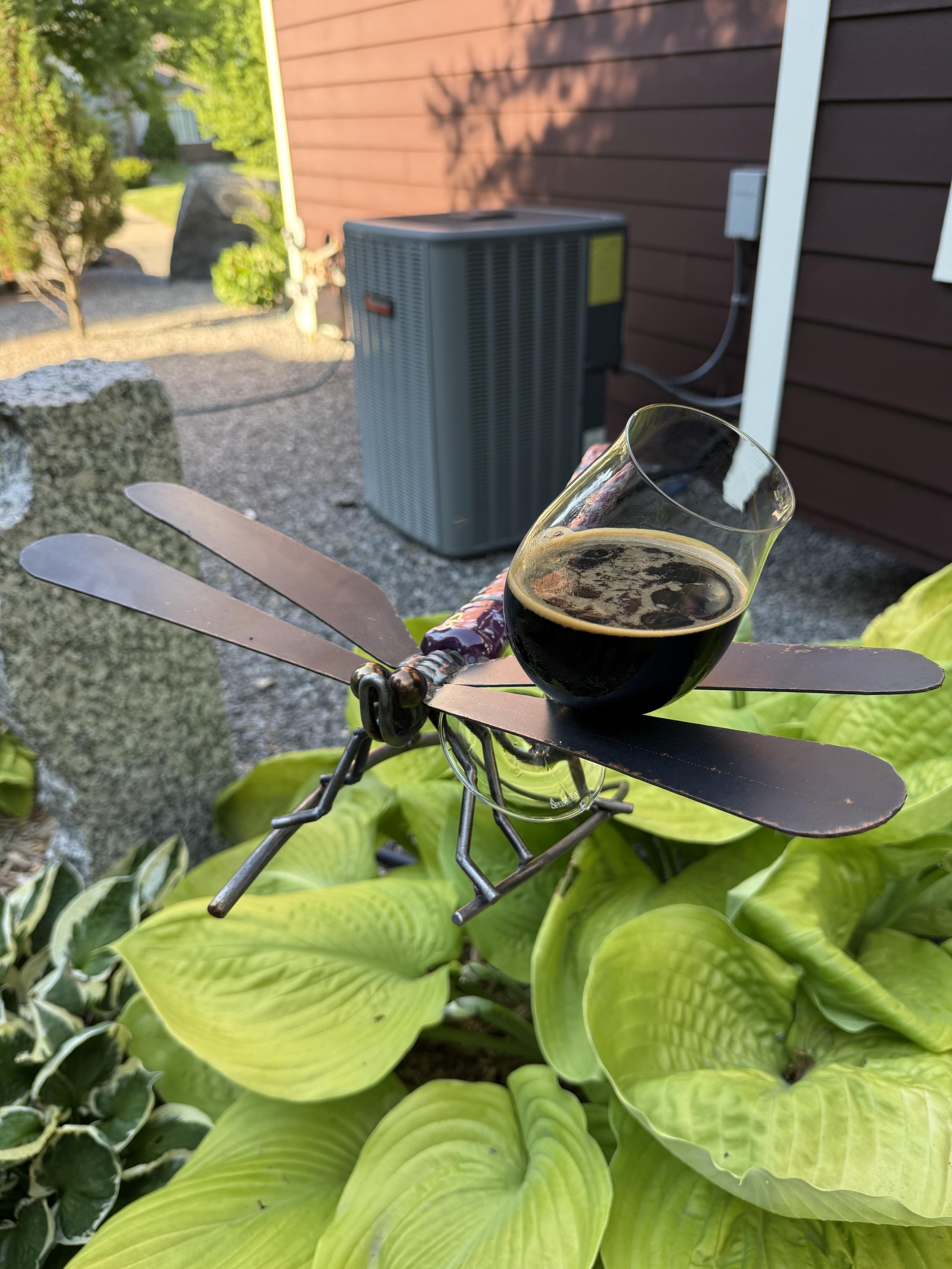 A large metal dragonfly absconds with a glass of beer above a giant green hosta plant. In the background is an air conditioner compressor unit.