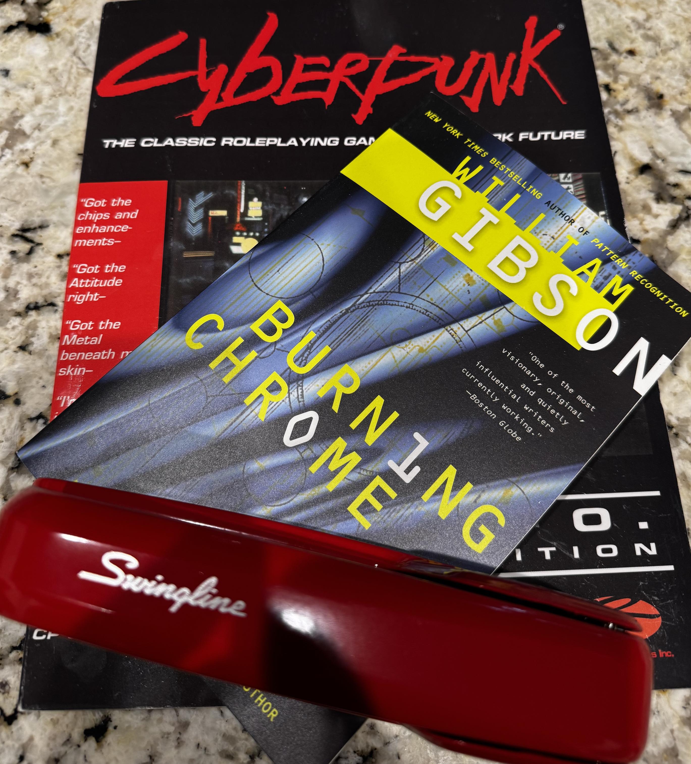 A picture of the Cyberpunk 2020 RPG rulebook, william Gibson’s “Burning Chrome” book, and a red Swingline stapler like that in the Office Space movie.