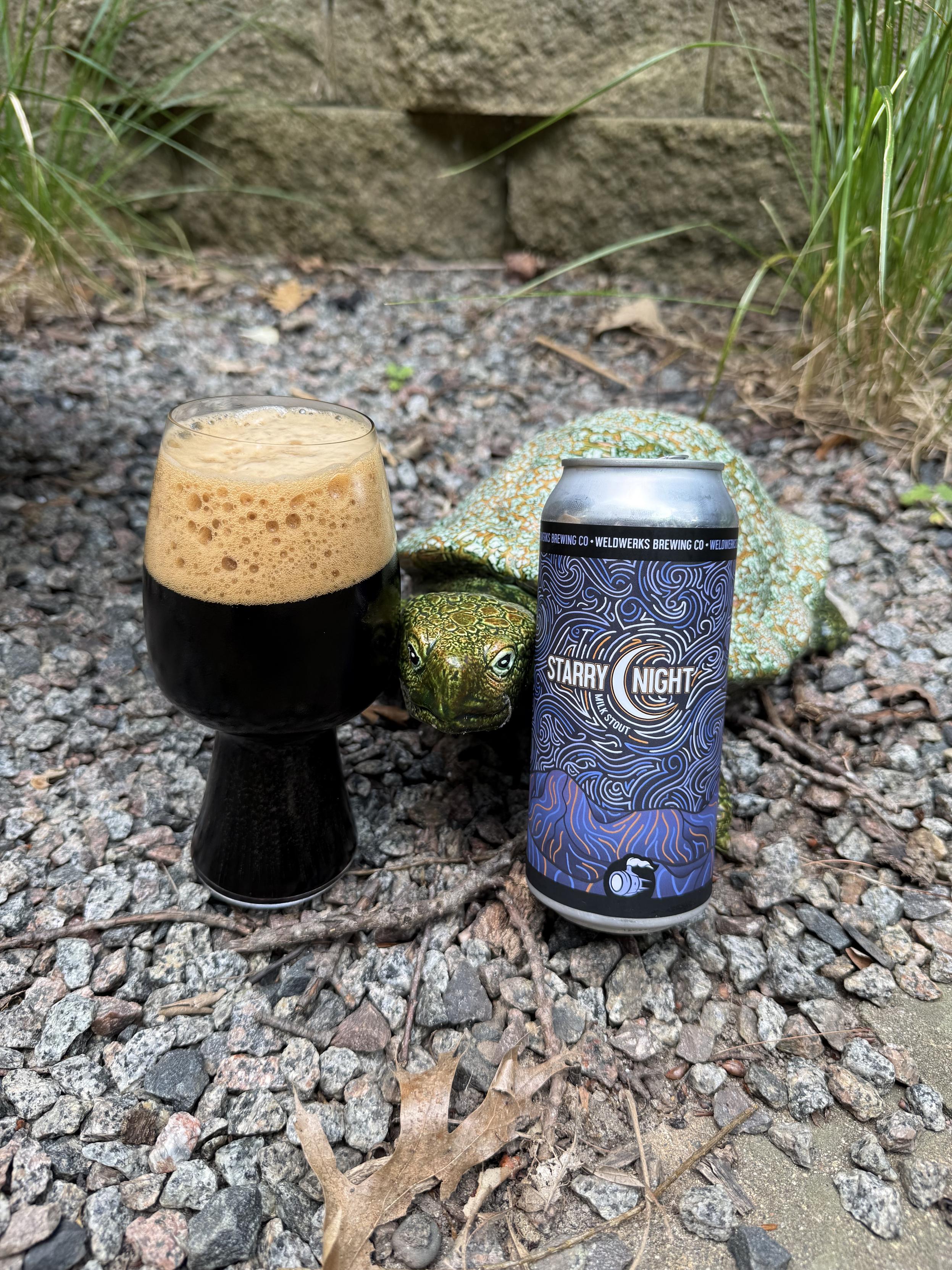 A stout glass full of dark beer with a tall bubbly tan head sits next to a large green turtle on a bed of crushed gravel. A pint can with a nighttime motif shares the view