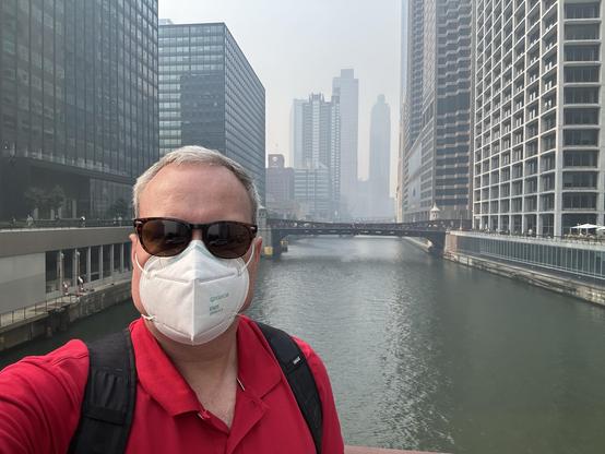 Man in sunglasses and an N95 mask taking a selfie with the Chicago River and tall buildings obscured by smoke in the background 