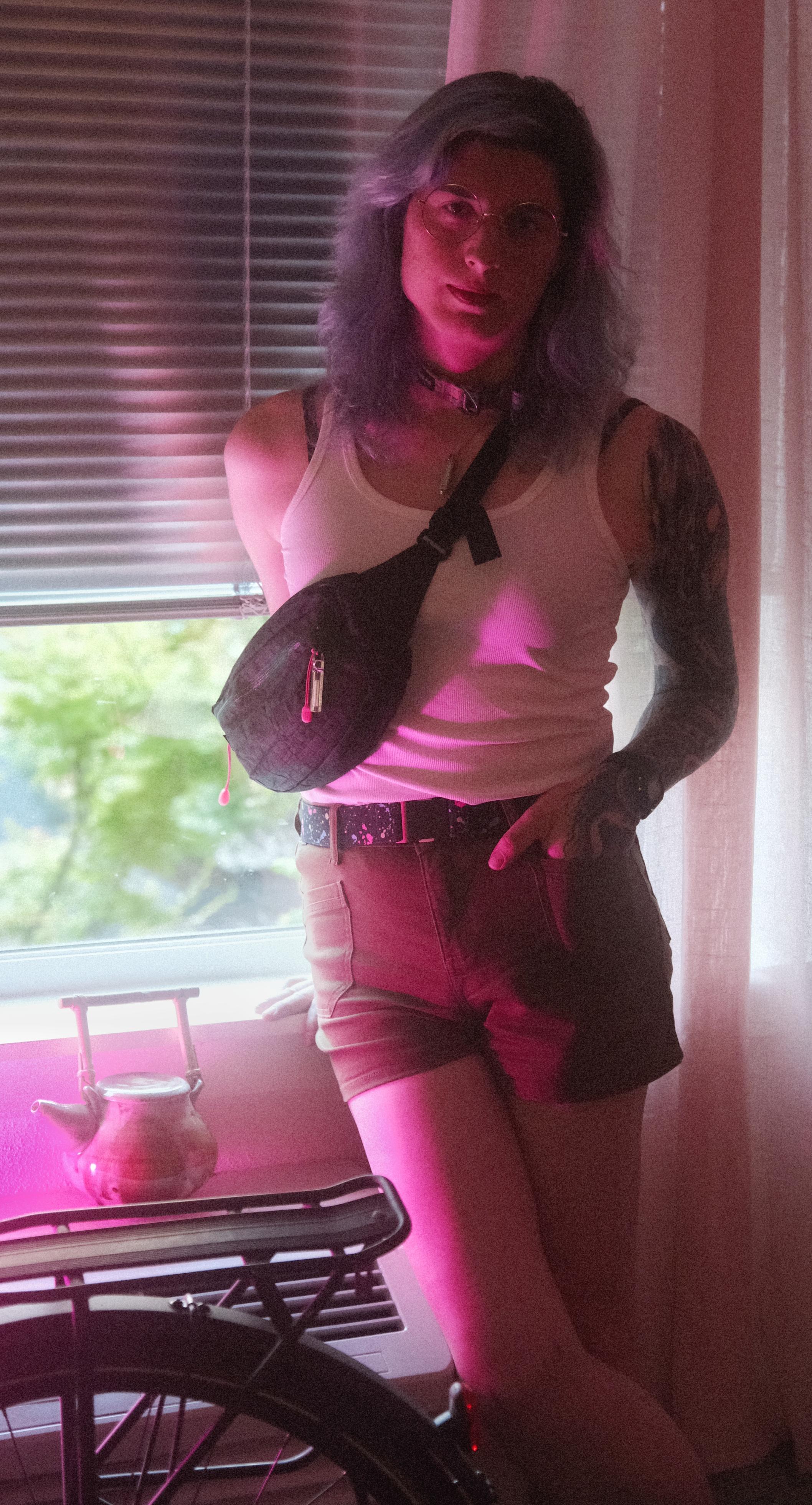 raven wearing a white tank and beige?? idk the color shorts in a pose leaning back against the window sill. her bike, a teapot she made are visible. there is a pink light just out frame