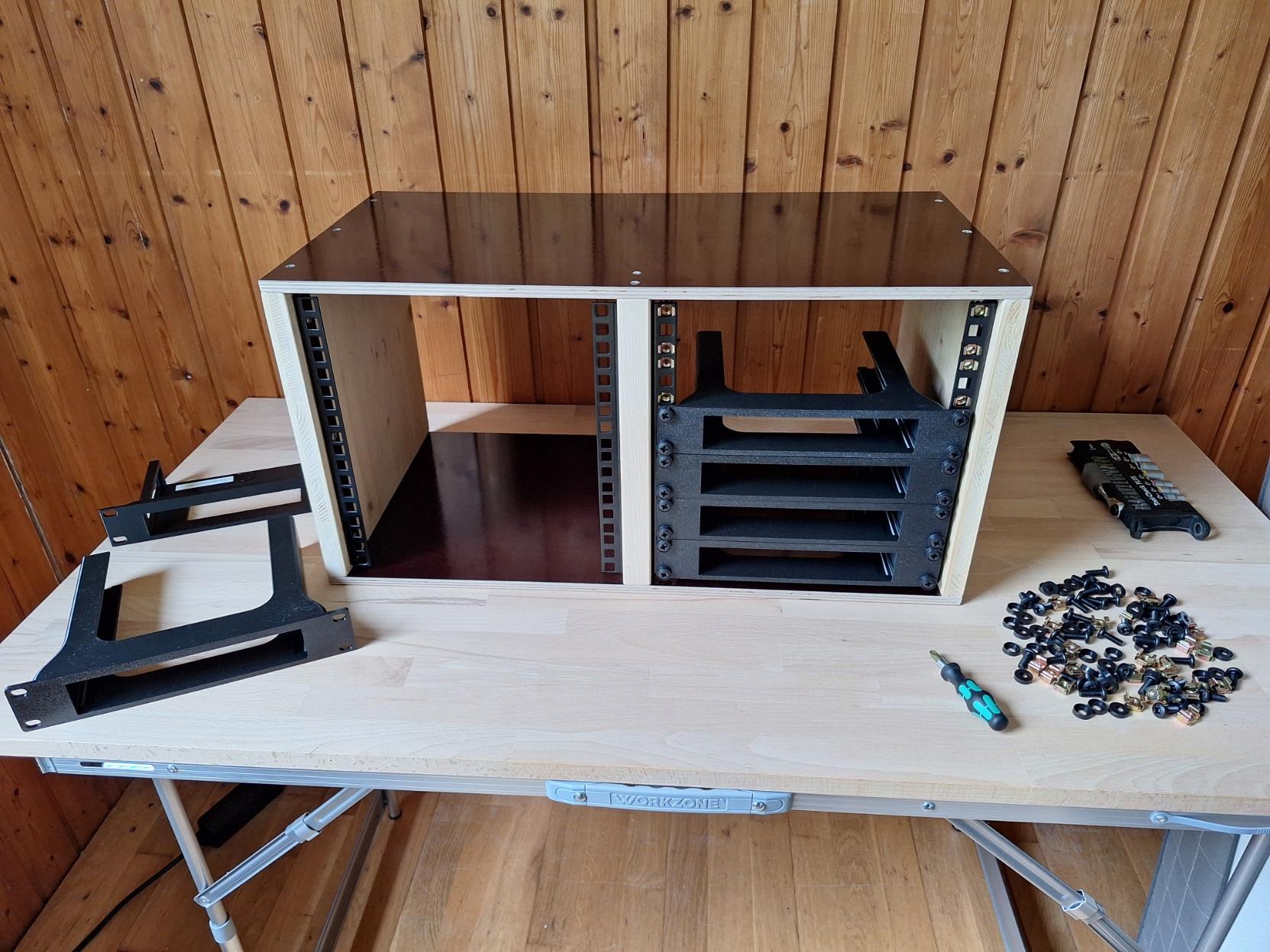 A wooden box with black rails on the sides and down the centre. Some plastic components are screwed onto the rails. Next to the box are screws, washers and rack nuts, as well as a screwdriver, a set of bits and two further plastic components.