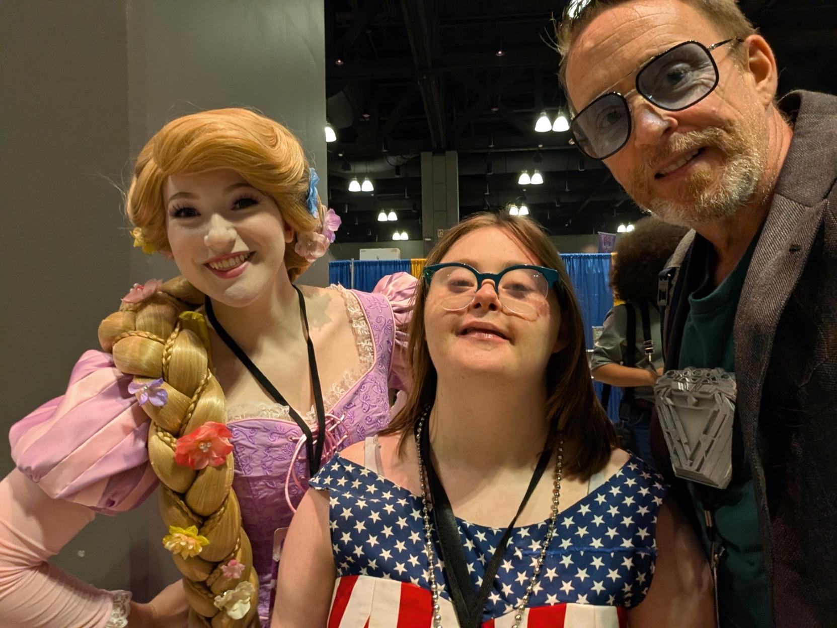 A woman with Down syndrome dressed as Captain America Princess between a woman cosplaying as Rapunzel from Tangled and a man cosplaying as Tony Stark