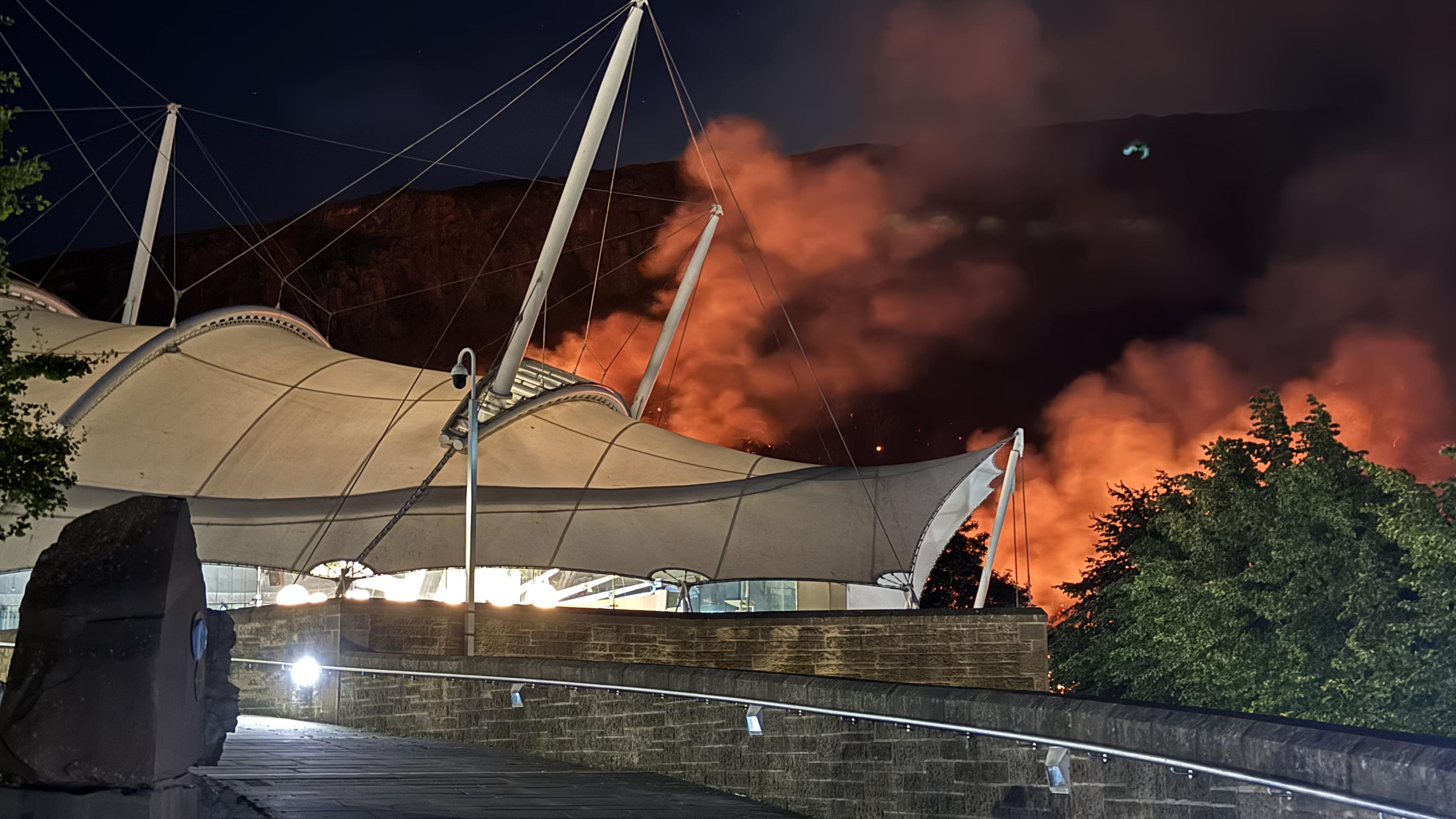 My Dynamic Earth, a tensioned fabric building lit up at night, behind it, clouds of smoke lit up orange by a fire, Salisbury Crags can be seen behind glowing orange against a dark blue night sky.