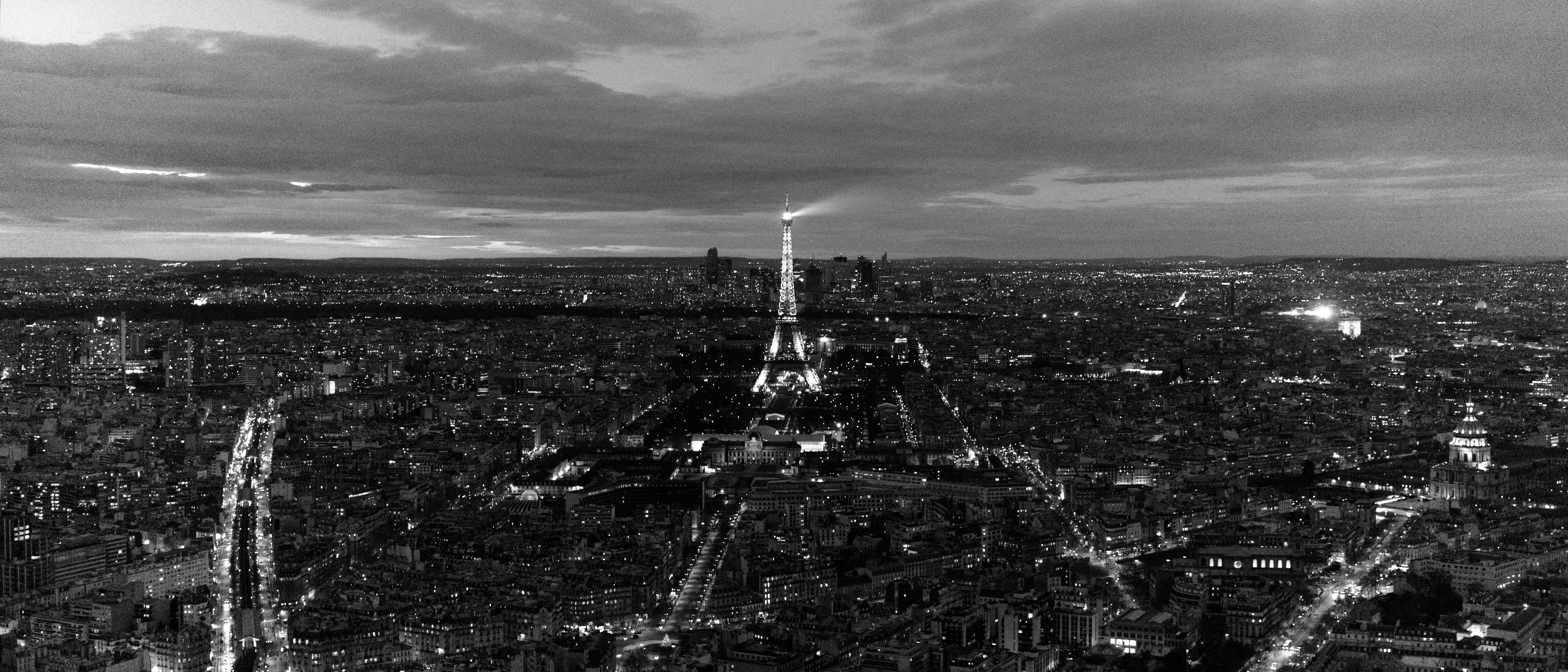A monochrome photo of the Eiffel Tower at dusk from the roof of a building. The sweeping light on the tower is visible as a light cone.