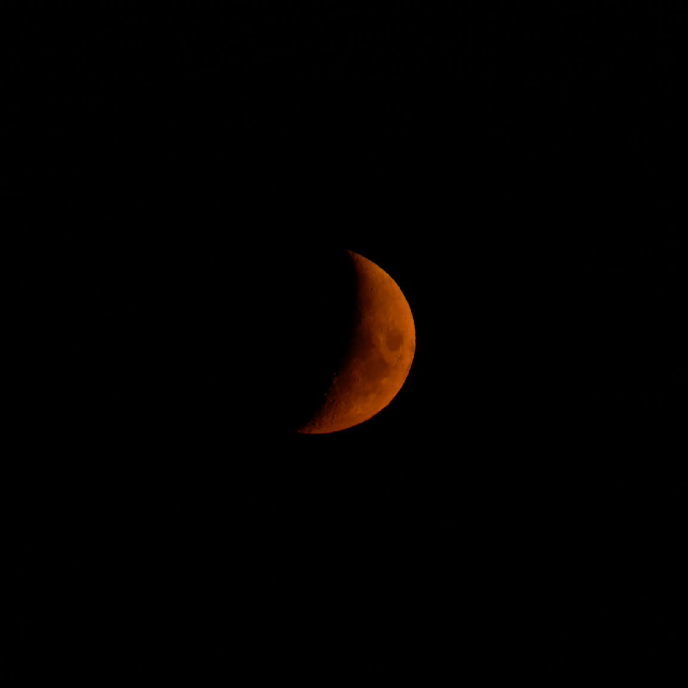 A crescent moon glowing deep orange-red against a pitch-black night sky, its cratered surface faintly visible along the illuminated edge.