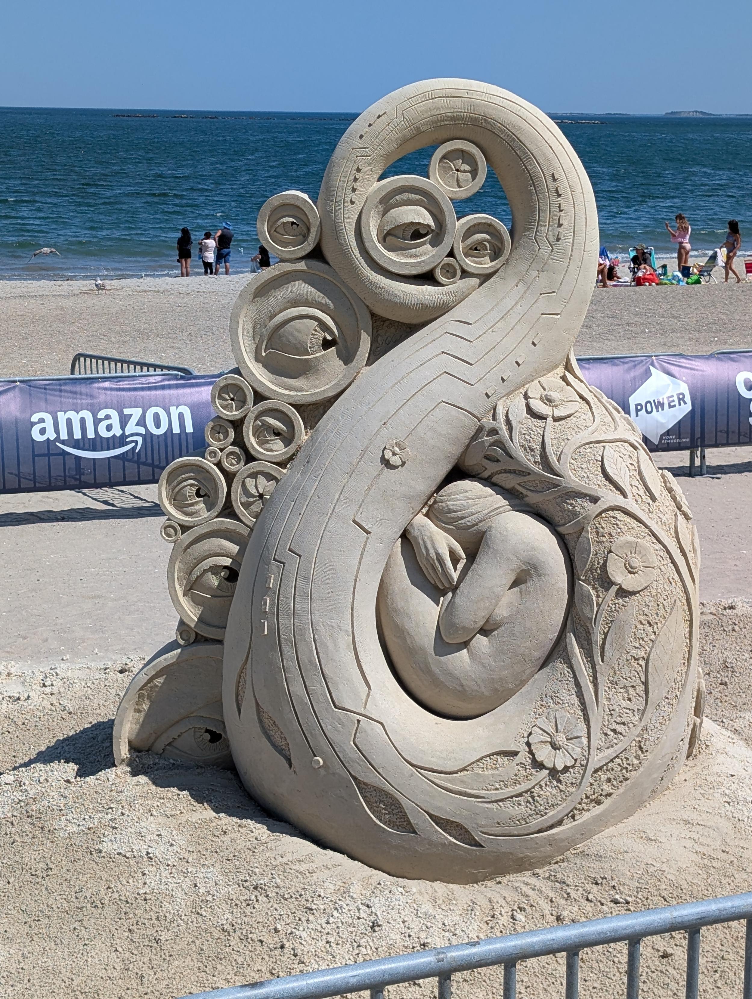 Photo of a sand sculpture entitled "Digital Hideout" by Hanneke Supply, featuring a female figure curled up inside a giant tentacle decorated with eyes.