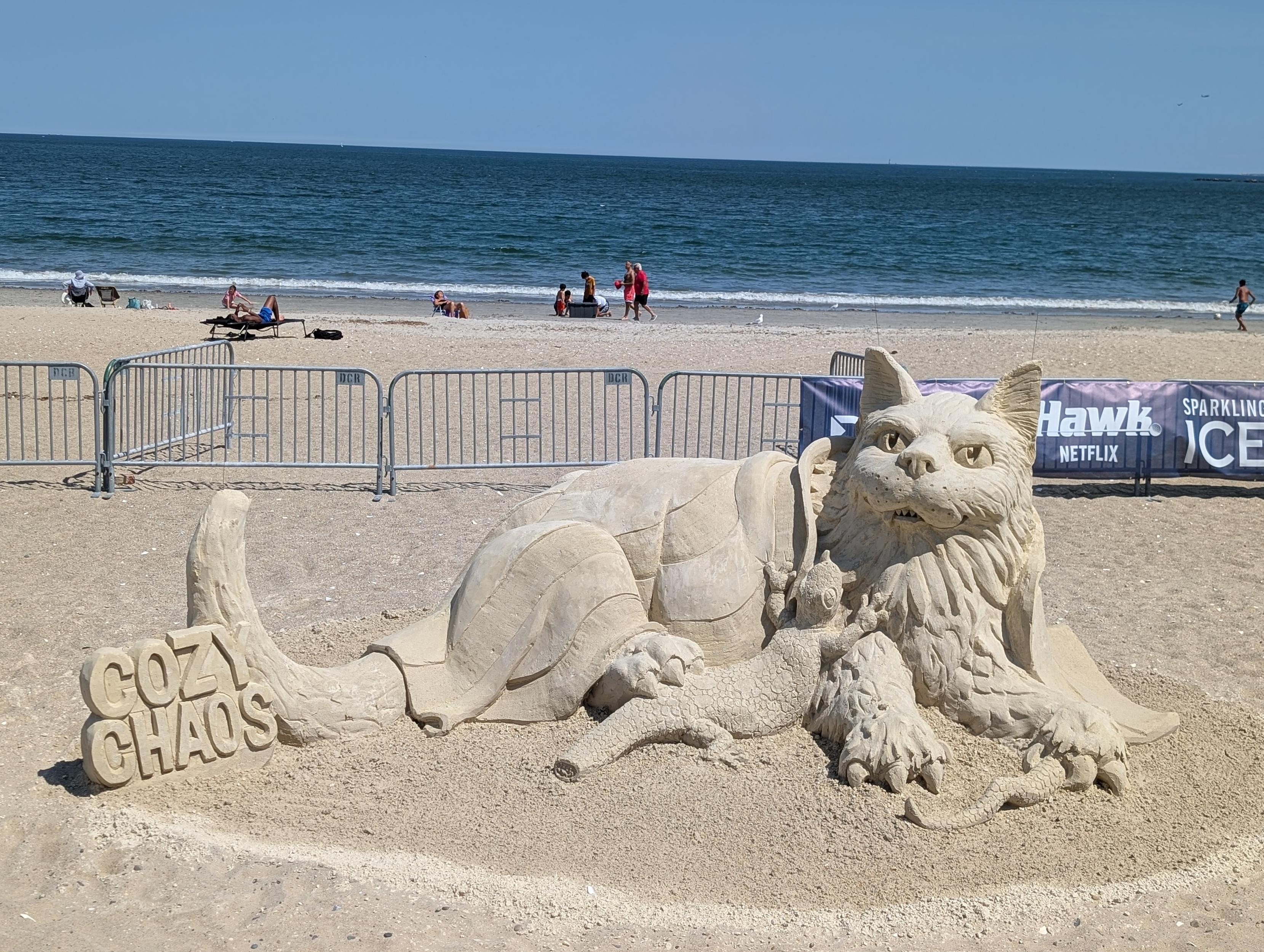 Photo of a sand sculpture, "Cozy Chaos" by Patrick Harsh, featuring a cat cuddling some lizards.