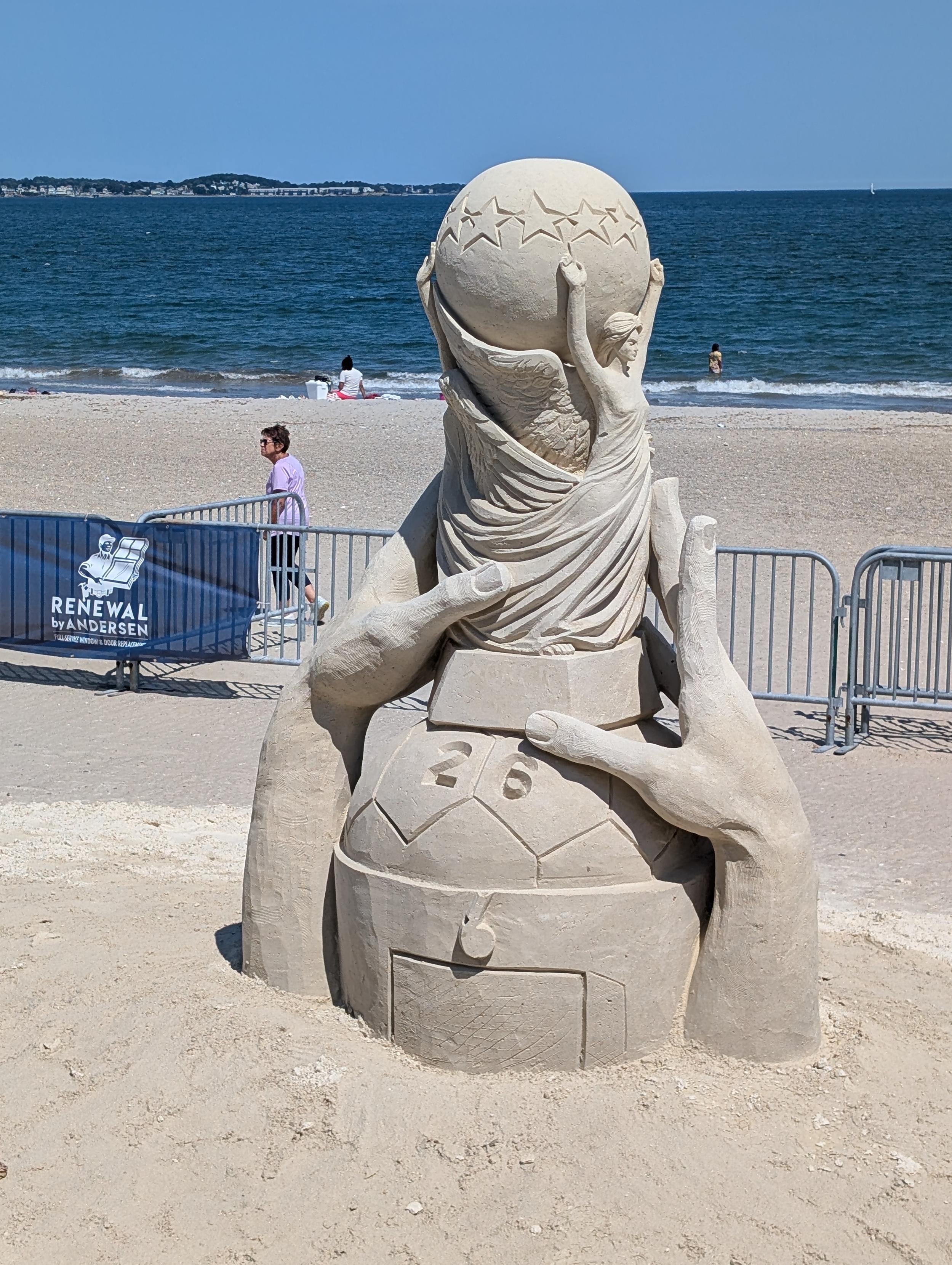 Photo of a sand sculpture, which features a replica of the 2026 World Cup trophy, being embraced by giant hands emerging from the beach.  (Sculptor unknown)