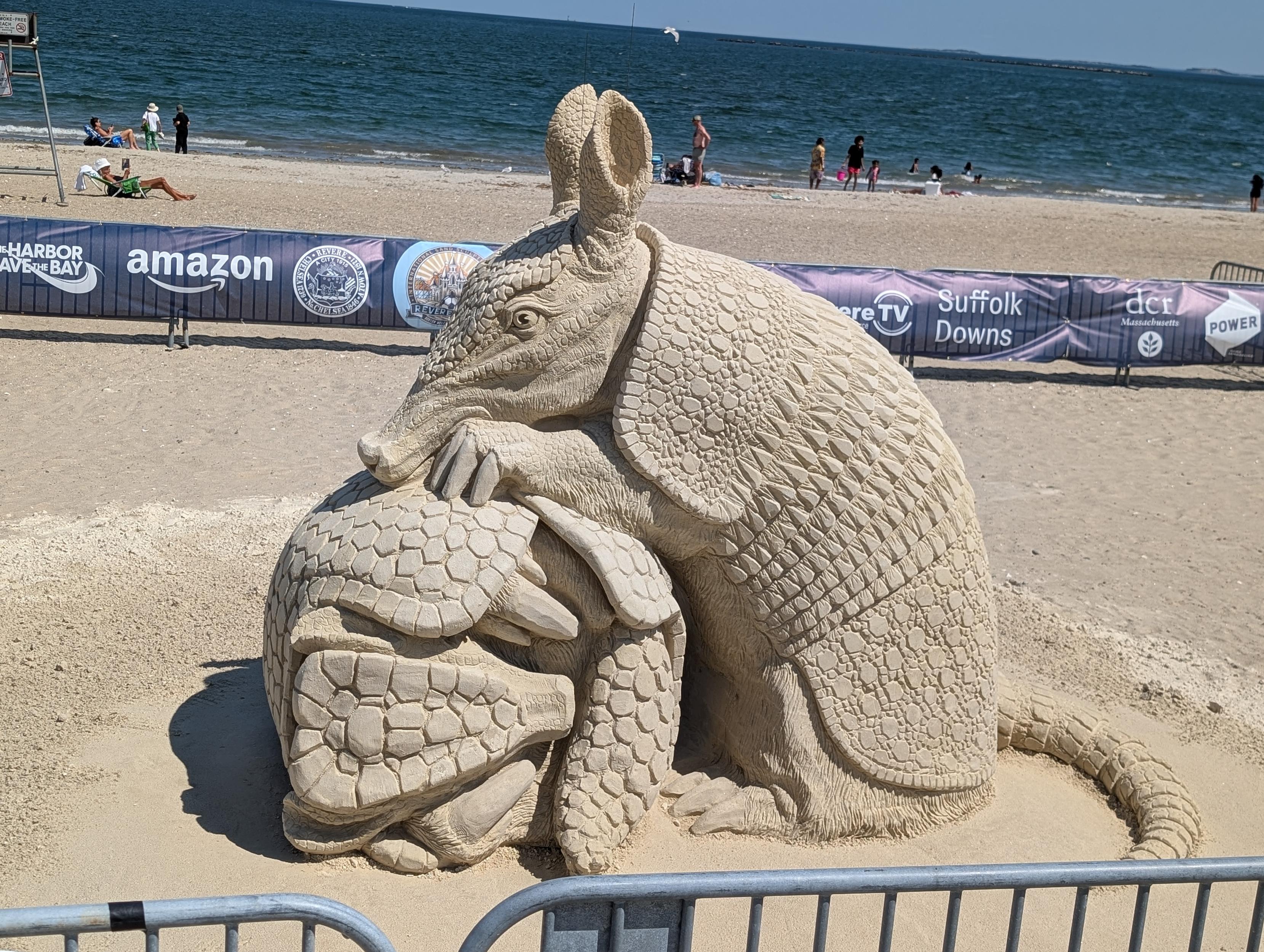 Photo of a sand sculpture, entitled "Overprotection" by Marie-Line Gagné, featuring one armadillo protectively resting atop another curled-up armadillo.