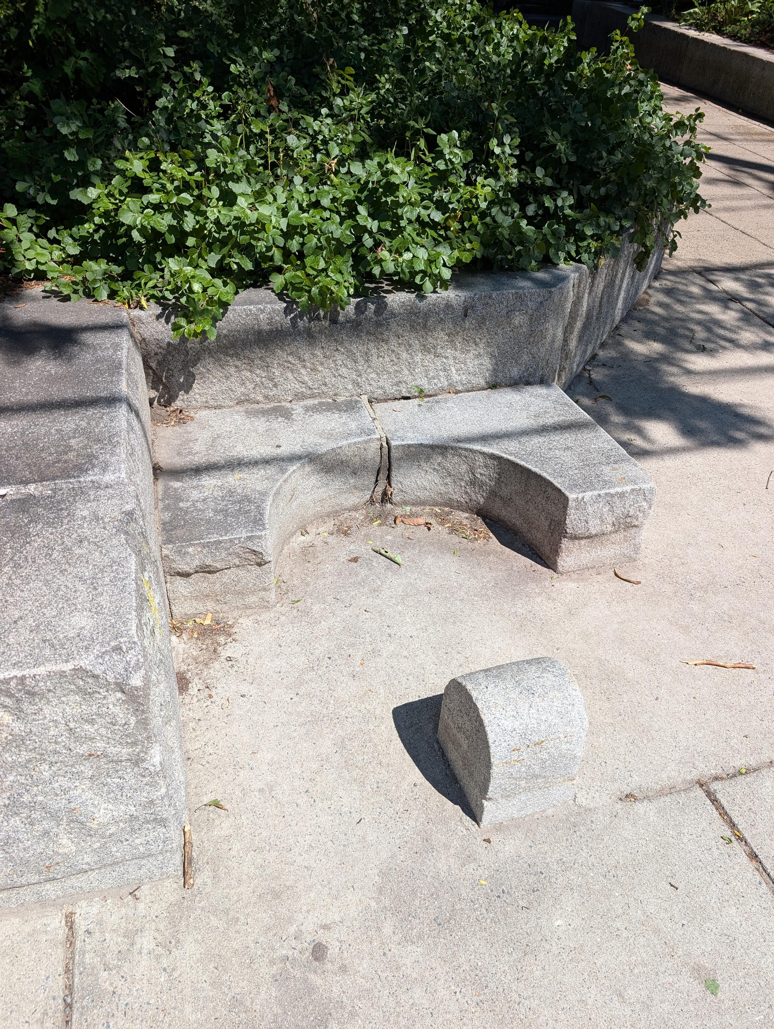 Photo of a foot-high stone semicircle at the base of a retaining wall, facing a small stone bollard.