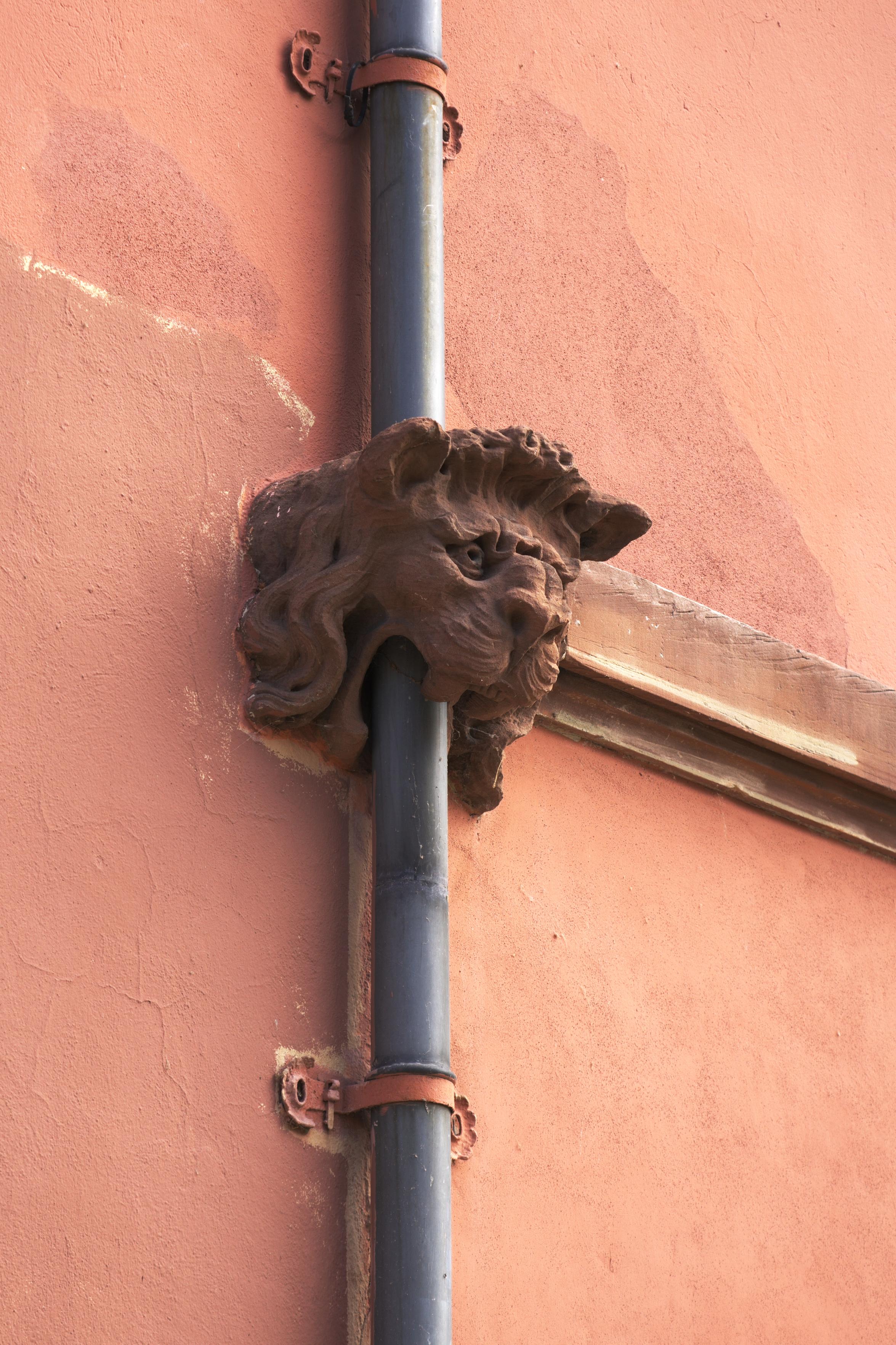 A red wall with a lion's head ornament (or waterspout?) through which a modern pipe has been shoved running down the wall.