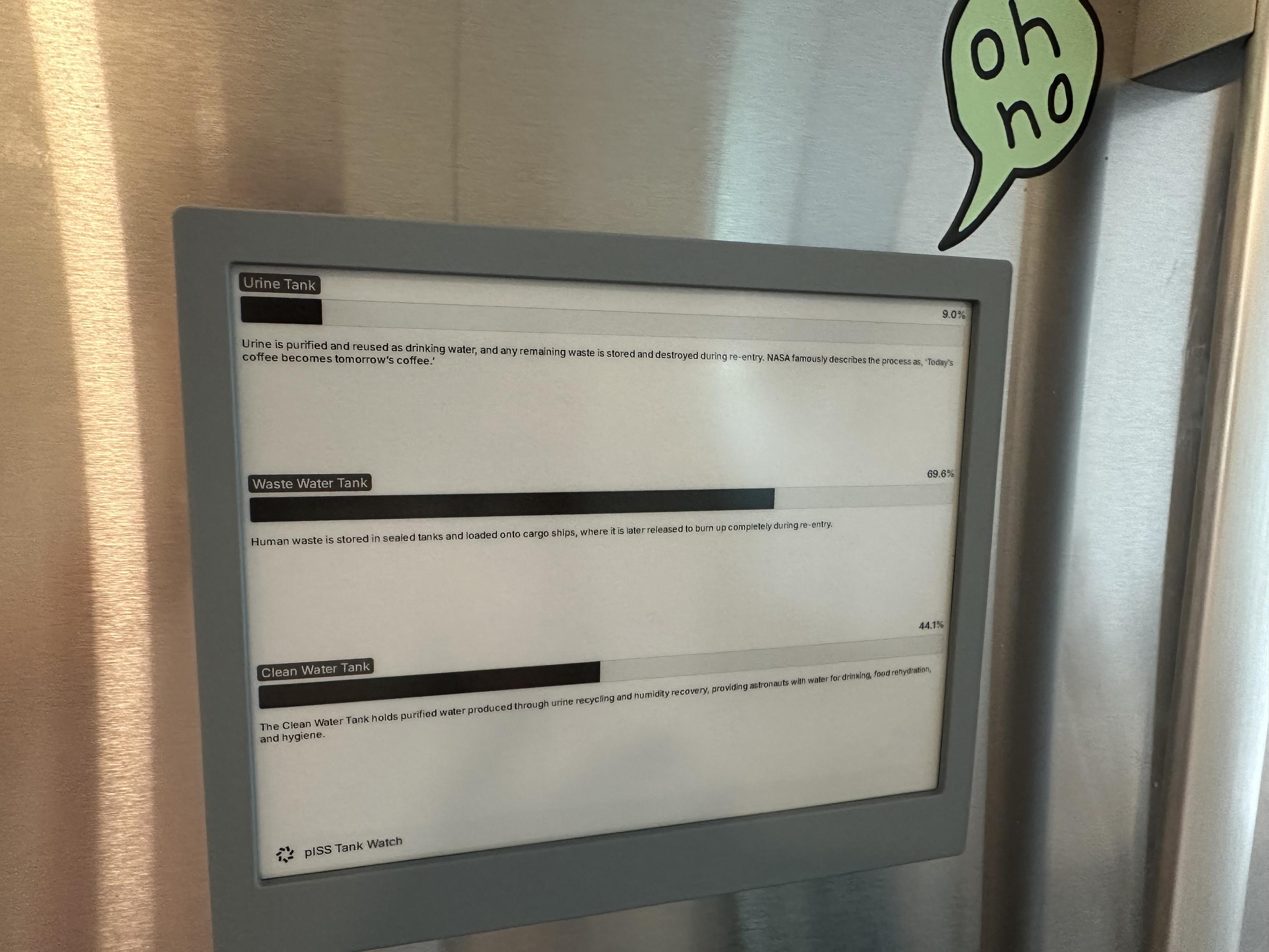 An e ink display on a fridge showing the levels of fresh water and piss onboard the ISS. 

There is a green fridge magnet that says “oh no” above it.
