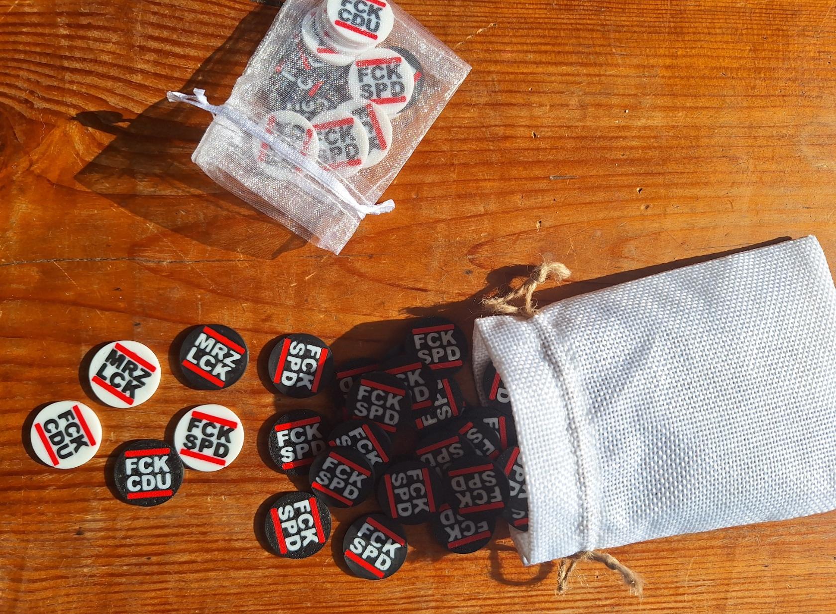 Zwei Beutel liegen auf einem Holztisch.
In den Beuteln und davor liegen einige runde Plastikchips in Grundfarbe schwarz oder weiß. Sie haben folgende Aufschrift, jeweils in der gegensätzlichen Farbe:

MRZ
LCK

FCK
CDU

FCK
SPD