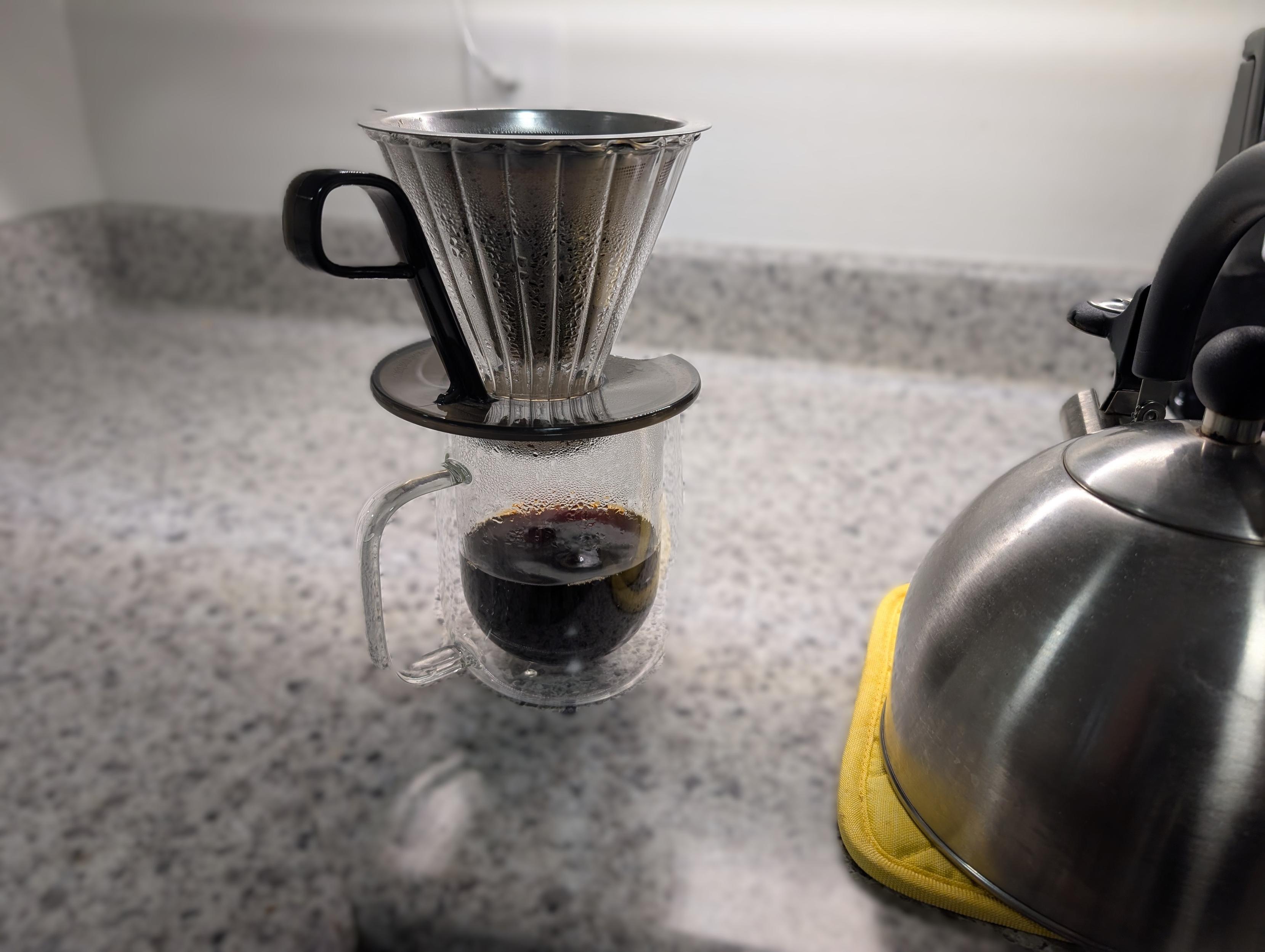 Clear glass cup on a white counter. On top of the cup is a glass and plastic filter holder. Inside the filter holder is a stainless steel, funnel-shaped coffee filter. The cup is about half full of coffee. To the right of the cup and filter is a stainless steel whistling tea kettle, sitting on a yellow hot pad.