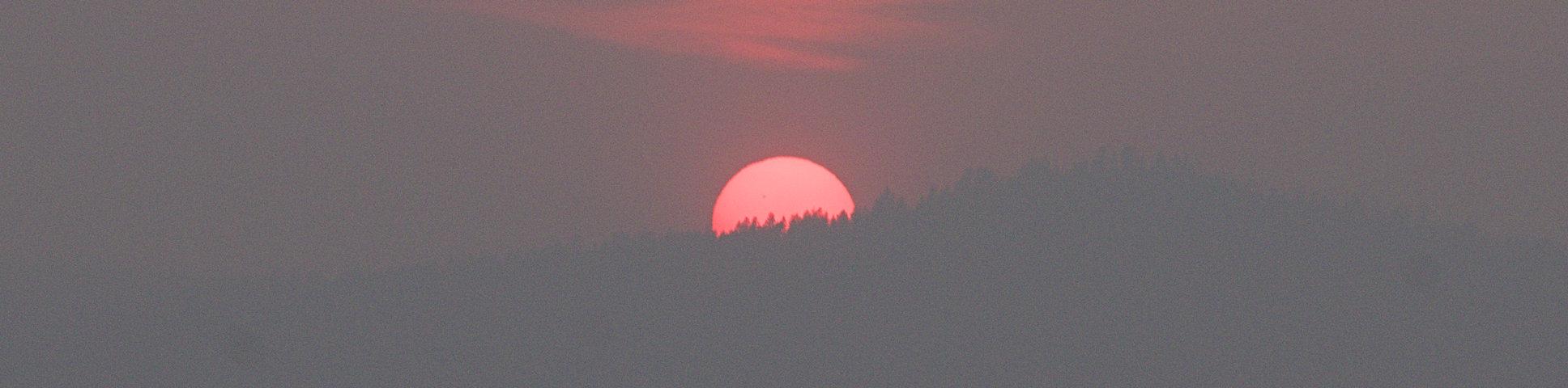A photograph of the sun setting below a tree covered mountain. The sun is very red due to smoke and a black spot is visible to the upper left of the sun's disk.
