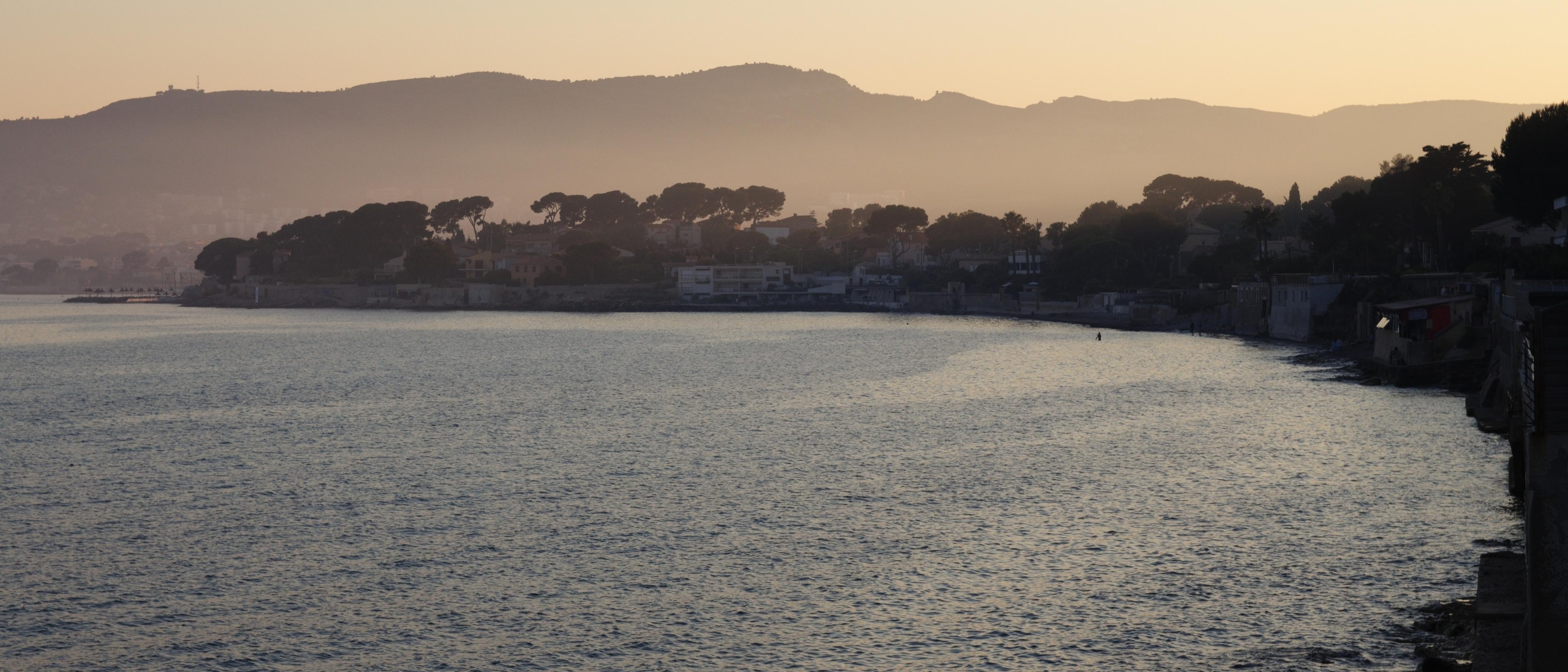 A morning photo of a crested bay with houses on the waterfront. A tiny person can be seen in the water.