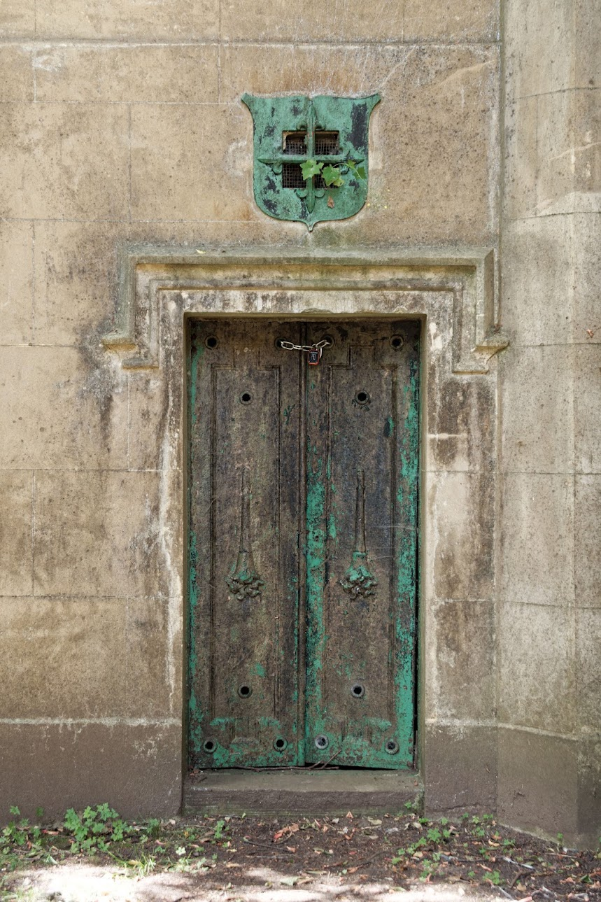 A weathered, dark metal double door with chipped green paint and a small padlock chain secured at the top, set into a rough stone wall. Above the doorway is an ornate, shield-shaped green metal vent with a cross design and a small ivy sprig growing through it.