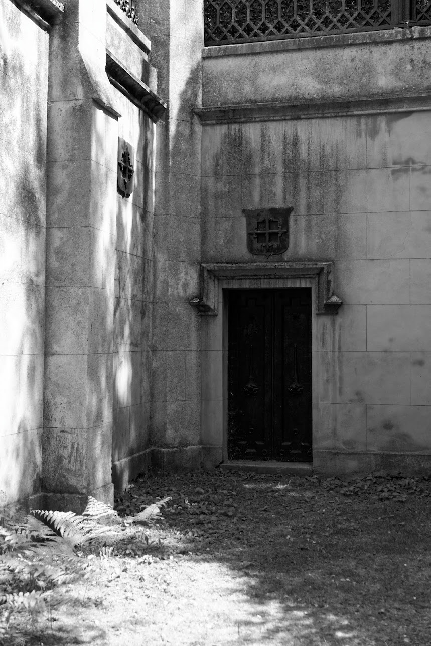 A black and white photograph of a dark, recessed double door set within a weathered stone wall corner, featuring a shield-shaped emblem above the frame, stone tile blocks, and sunlit ferns in the foreground casting dappled shadows.