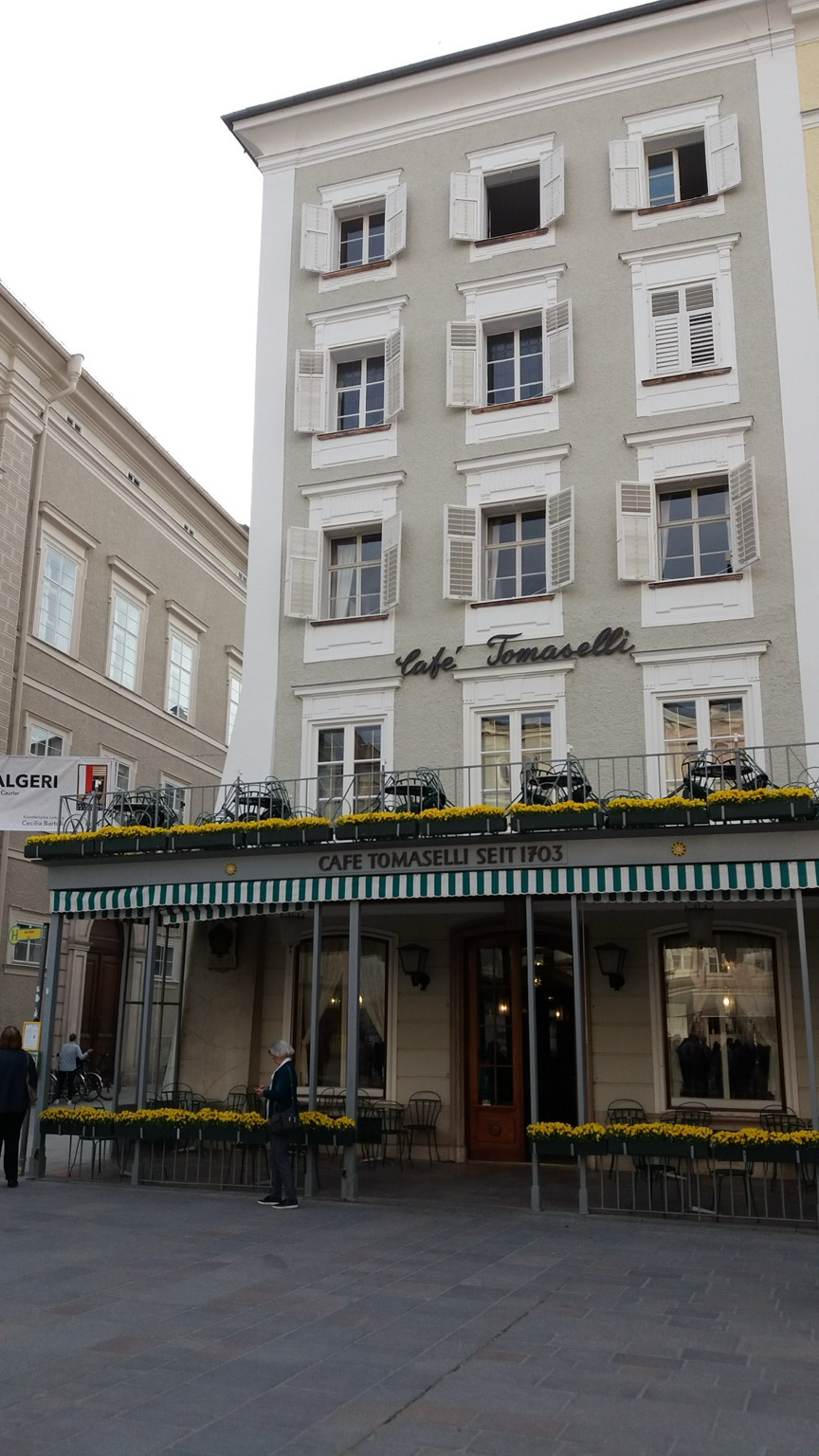 We are looking at Cafe Tomaselli, a hangout of Mozart in Salzburg, Austria. The five story building is grey with white trim. Green and white striped awning protects patrons entering.