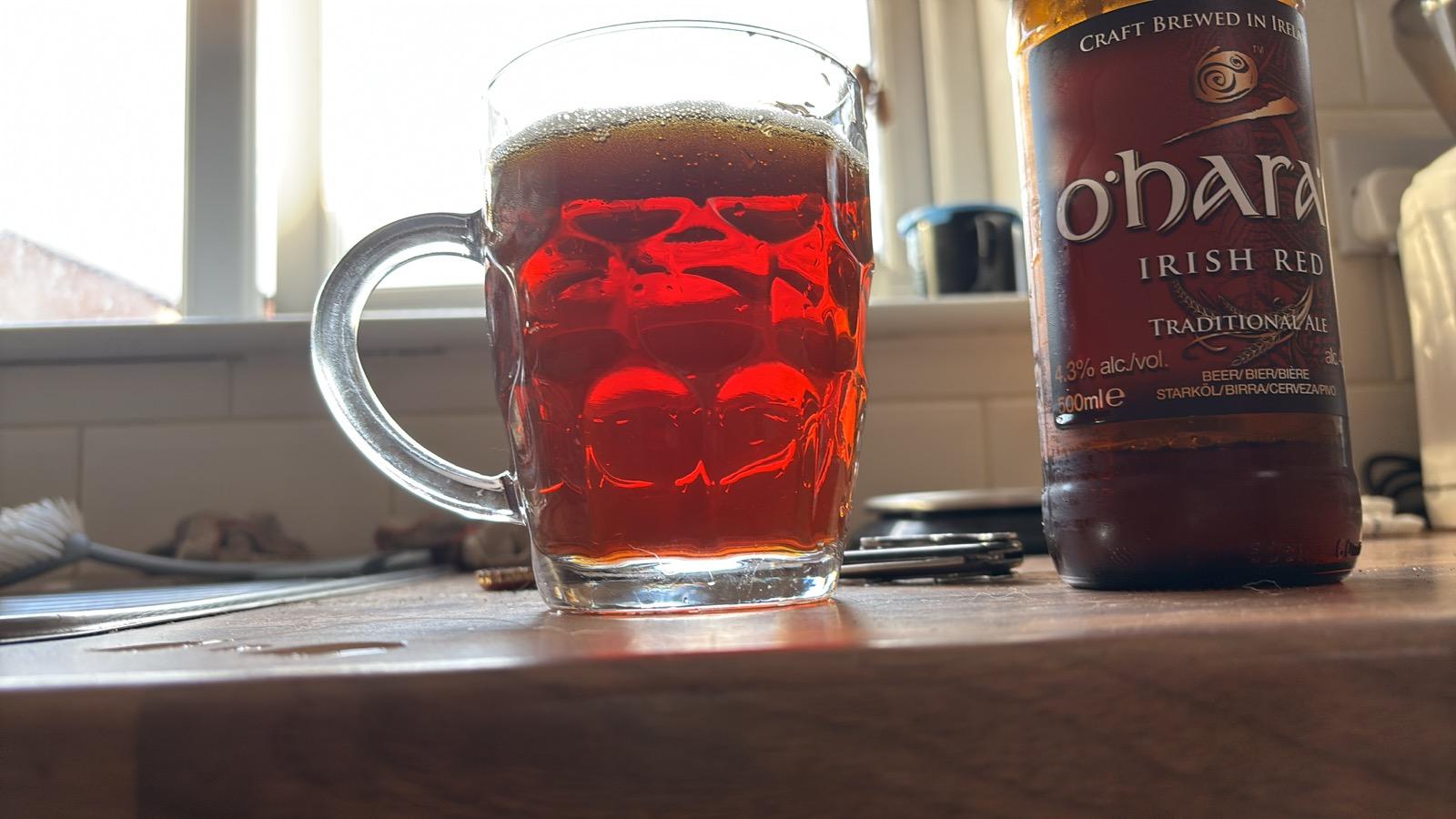 dimpled pint mug, backlit by a window, filled with a deep red beer. A bottle of O’Hara’s Irish red is to the right.