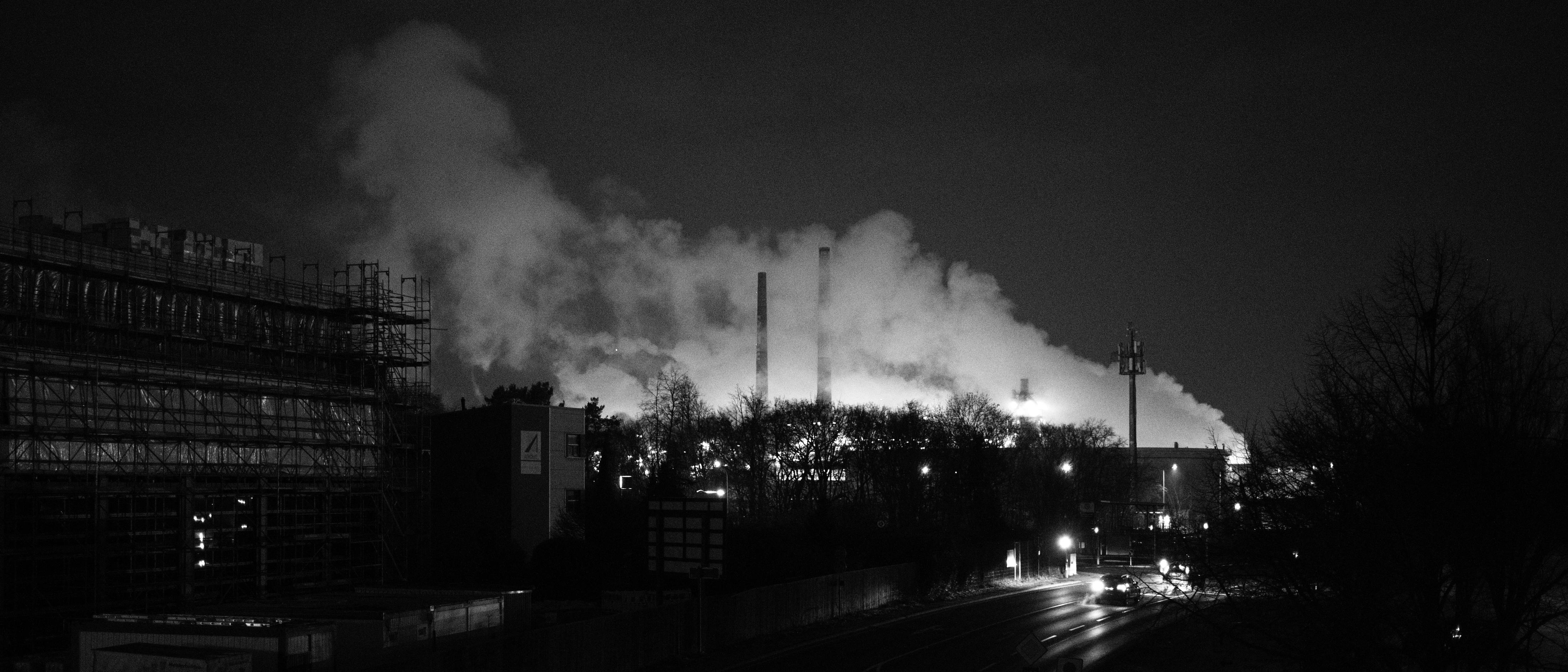 A black-and-white, nighttime photo. A brightly illuminated industrial complex in the background spits out billowing, white smoke and illuminates it from below.