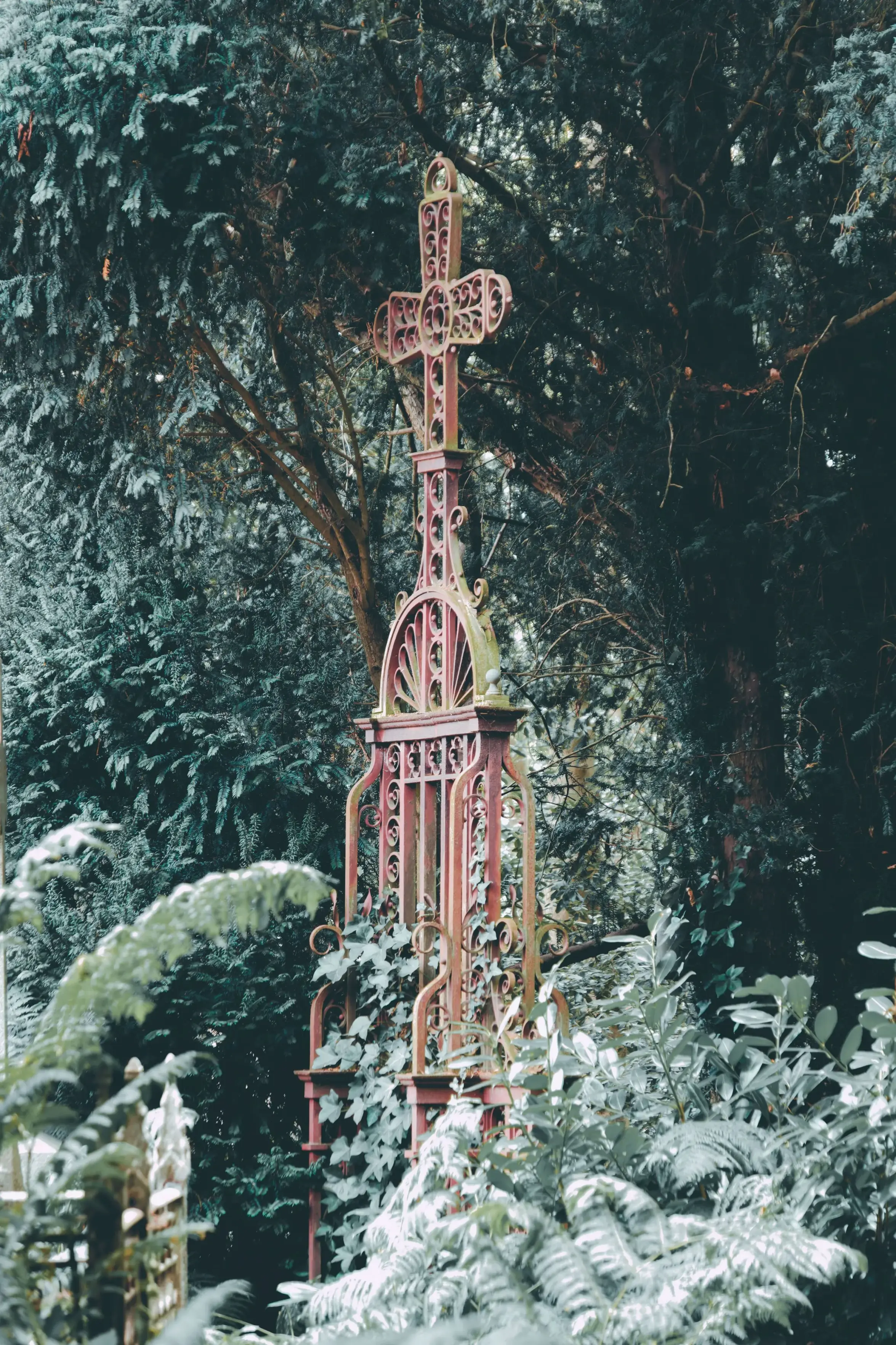 An ornate, weathered reddish-brown iron grave marker topped with a decorative cross stands vertically amidst dense, dark green foliage and ferns in a moody cemetery setting.