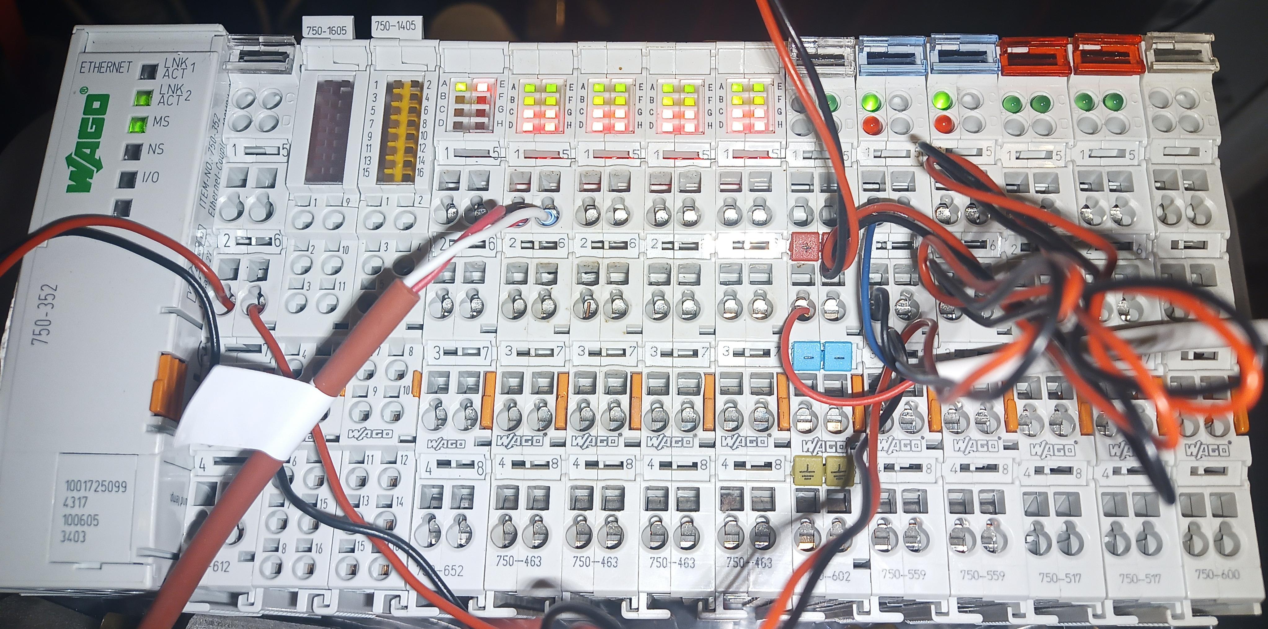 An industrial control I/O system, manufactured by Wago, as observable by the logo everywhere. The system is modular, about a dozen individual modules are visible. They are white, narrow, but tall, and fit snugly side by side like lego bricks. Besides a couple of LEDs, the front is covered by the typical Wago spring connector system: a round hole for the wire, a square hole for the tool that operates the spring holding the wire. A few wires already connected gives a sense of scale: it's about the width of a hand vertically, and the length of a hand vertically.