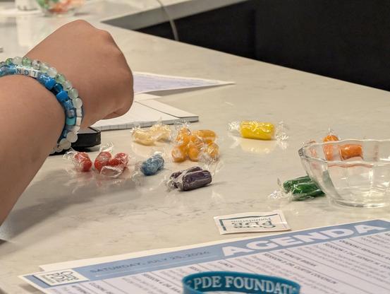 An image shows an arm wearing a blue, white, and clear beaded bracelet resting on a white surface next to scattered candies and a document. Various individually wrapped candies in colors such as red, yellow, orange, orange, blue, purple, and green are spread across the center of the table, with some pieces inside a small clear glass bowl on the right. A printed paper at the bottom contains text including "SATURDAY, JULY 25, 2026", "AGENDA", "LUNCH", and "PDE FOUNDATION". Partially visible text …