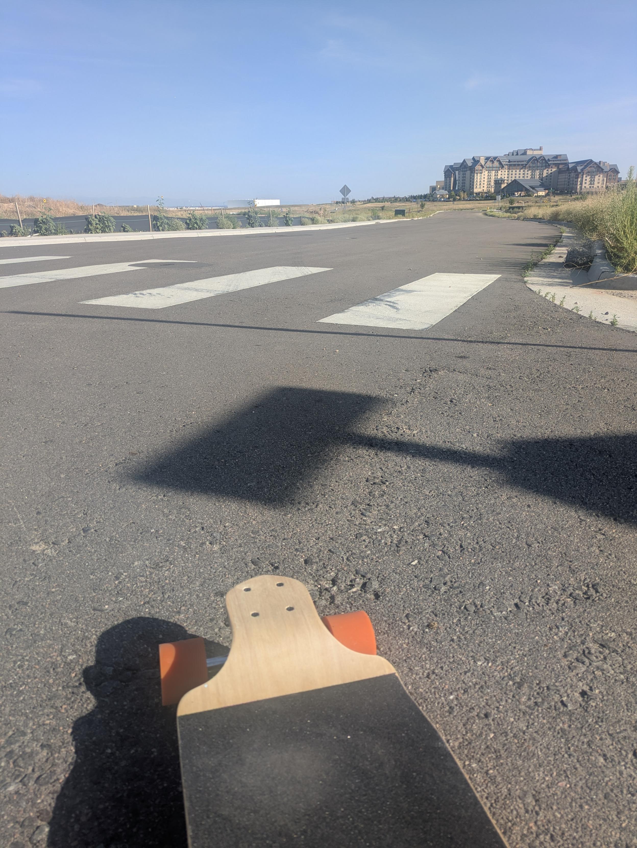 Picture of the nose of a longboard pointing down a street with a nice down hill grade. No cars on the street, no buildings either. Clear blue sky's.