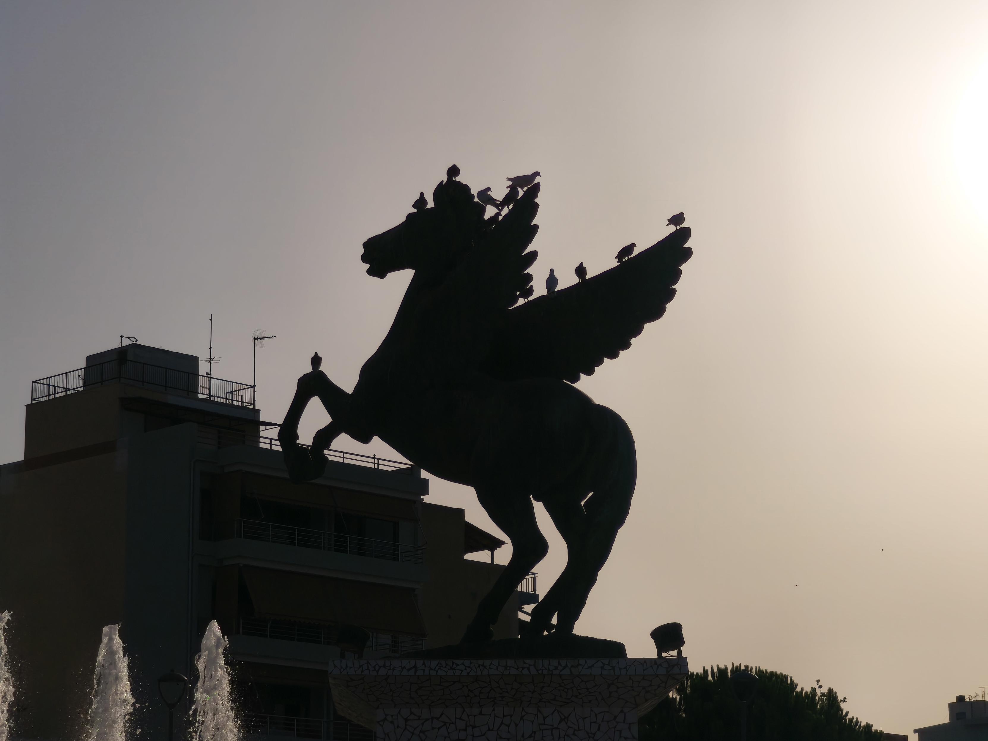 Picture of the statue of Pegasus in Corinth, Greece. Pigeons are sitting on the statue. The image itself has a dark contrast due to the sun.