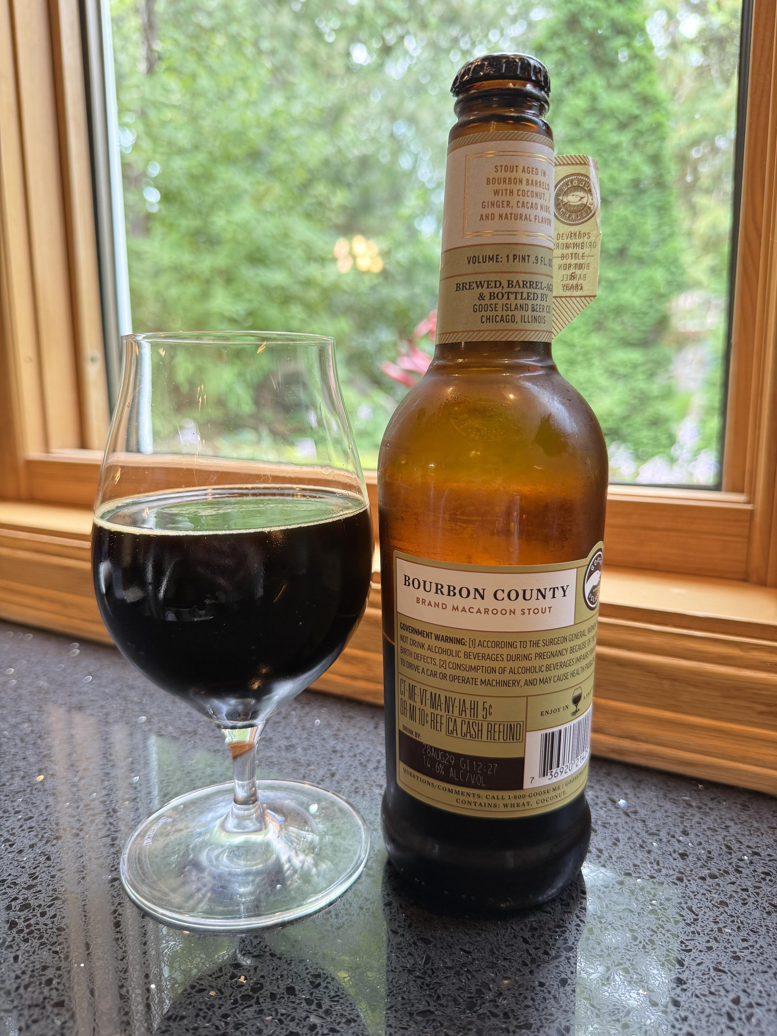 A snifter glass of dark beer is next to a tall brown bottle with a tan and darker tan neck label and body label. It’s another bright sunny day outside the window