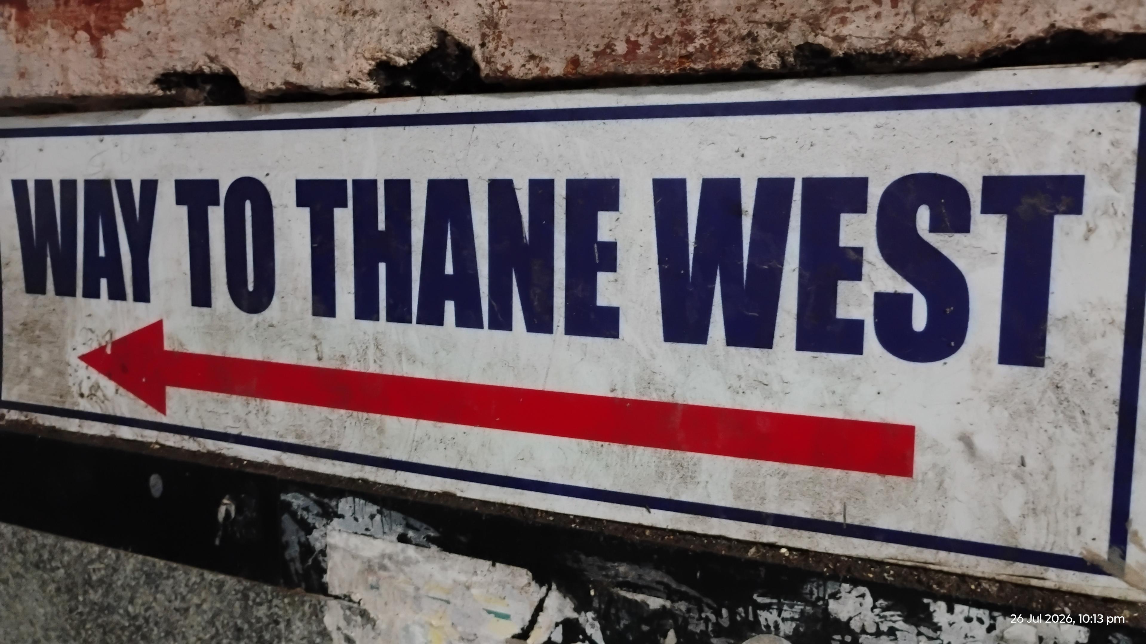 A board in a railway station that reads "way to thane west" in blue letters with red arrow below it pointing left.

The board is placed on a very damaged and/or inconsistent wall