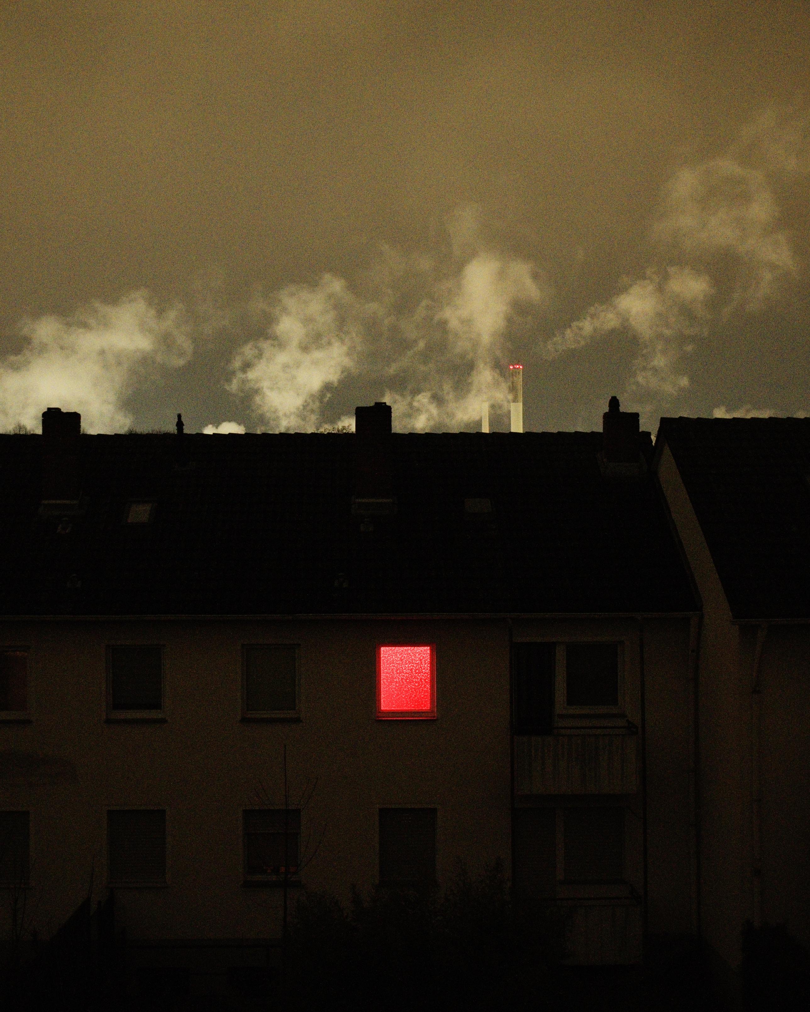 A nighttime photo of a house with a single illuminated window glowing in red. The image is grainy. The background is illuminated by light from a factory, and smoke rises to the sky.