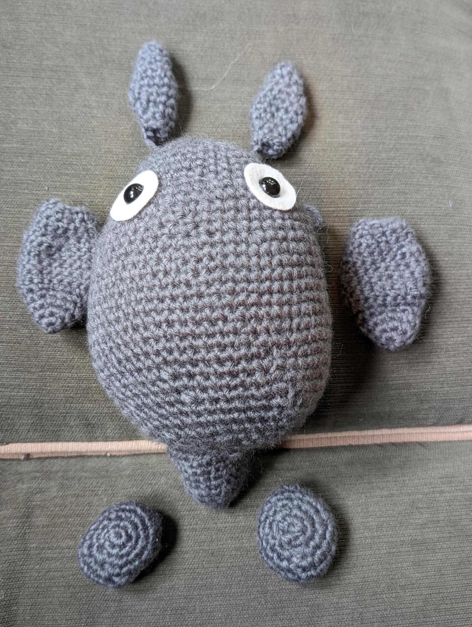 a grey crochet Totoro doll body with eyes, but no belly, surrounded by unattached ears, arms, feet, and tail