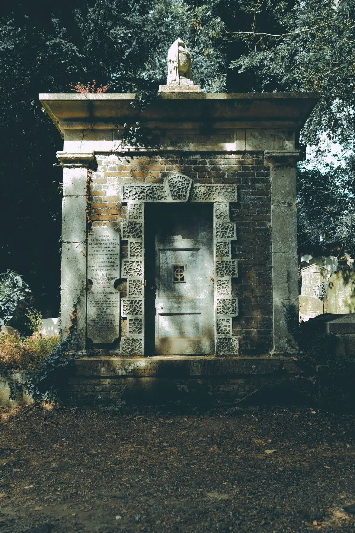 A weathered brick mausoleum set within a shaded cemetery features a central metal door surrounded by an ornate, textured stone frame with intricate patterns and a small barred window with a cross design. A carved stone urn rests on the flat roofline, and an inscribed memorial plaque is mounted on the stone pillar to the left of the entrance. The structure is flanked by heavy stone pilasters and overgrown foliage, with dappled sunlight filtering through the dense trees above.