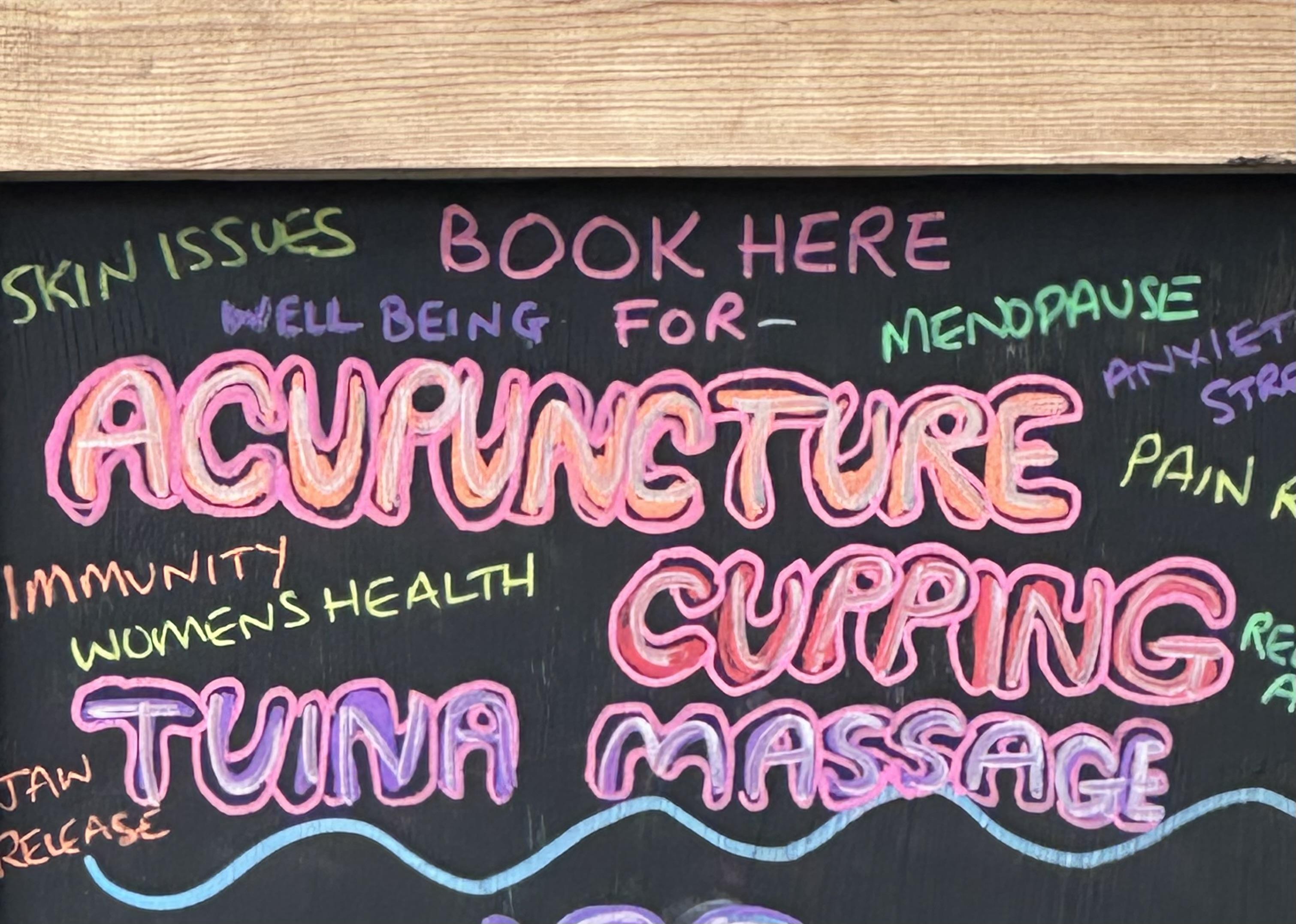 A chalkboard advertising sign in many colours and words. Amongst it it says Tuina Massage but the kerning makes it look like Tuna.
