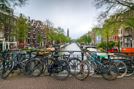 A scenic view of cannal with boats and 5 story apartment homes. with Bicycles arcross a brick bridge and a tower in the distance. very lovely and inviting setting.