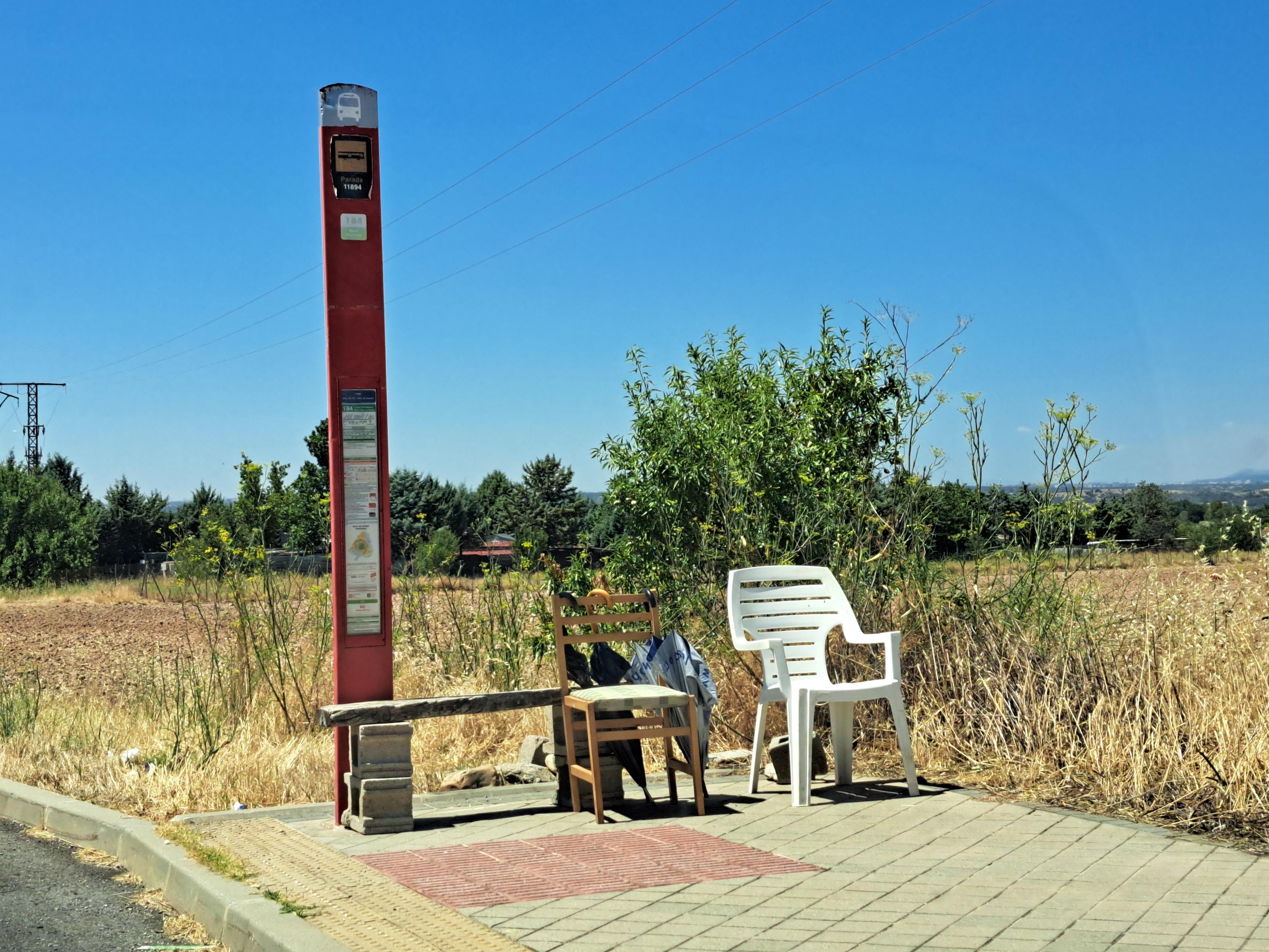 En la fotografía se ve un poste indicando el número de parada y la única línea que pasa por ahí, la del 184.

Pegado al poste rojo hay un tablón de madera sustentado por unos bloques de hormigón para  sentarse.

Al lado del improvisado banco, hay 2 sillas, una de madera y otra de plástico.

En el respaldo de la silla de madera, hay 3 paraguas colgados para que los usuarios del transporte público se protejan del sol, ya que no hay ninguna sombra.