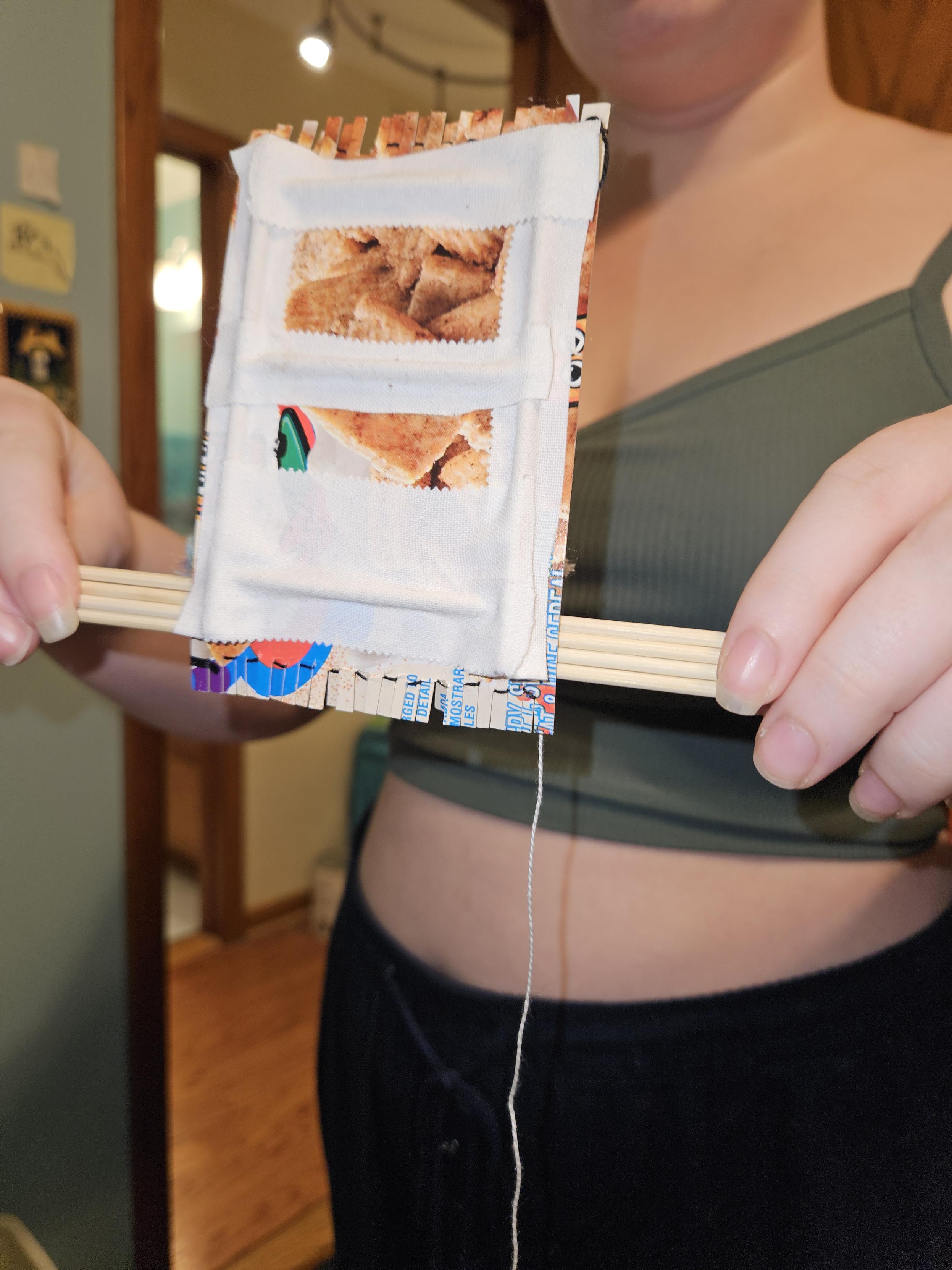 Underside of a hand-made mini-loom.  The base is a piece of cardboard from a cereal box, reinforced by some long bamboo sticks fixed to the cardboard with medical tape.  The long loom base is four bamboo sticks taled together.