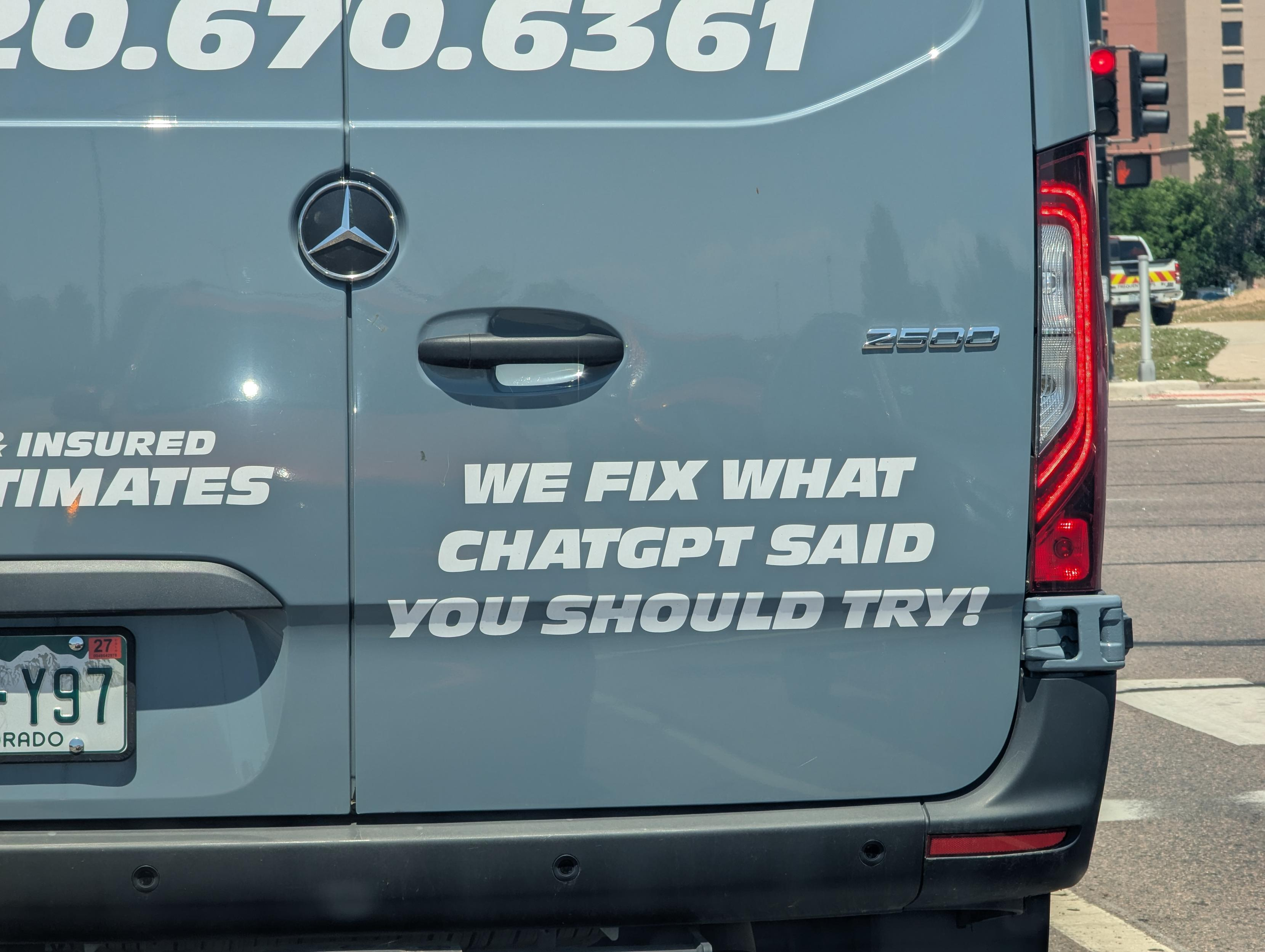 The image shows the rear of a gray commercial van, likely a Mercedes-Benz Sprinter, given the distinctive three-pointed star emblem visible on the left side of the van's rear door. The van has a smooth, matte gray finish that reflects some ambient light, creating subtle bright spots. Dominating the center of the rear doors are large, white, sans-serif text graphics. The main message reads "WE FIX WHAT CHATGPT SAID YOU SHOULD TRY!". Above this, to the left, are smaller white text elements, partially obscured, reading "INSURED ULTIMATES". To the far left, a large, white phone number, "20.670.6361", is printed across the top of the van. To the right of the Mercedes emblem, the model number "2500" is debossed in silver lettering.

The composition focuses tightly on the rear of the van, cutting off the top and sides of the vehicle. The perspective is slightly from the right and low, looking up at the rear doors. The lighting suggests a sunny day, with harsh overhead light creating reflections on the van's surface and casting subtle shadows.

On the lower left of the van is a license plate, which is partially visible. It appears to be a Colorado license plate with the state name "COLORADO" at the bottom and a plate number that includes "Y97". A smaller sticker or decal on the plate shows the number "27" and a small depiction of a mountain range. The rear of the van has a dark gray bumper with visible circular parking sensors. The taillights are integrated into the vertical sides of the rear, featuring a distinctive red LED pattern that is partially illuminated, suggesting the brake lights may be on or the vehicle is in a setting where they are active. A silver handle for the rear doors is positioned centrally above the main text graphic.

In the background, slightly out of focus, we see elements of an urban or suburban environment. To the right, a traffic light is visible with red and yellow lights illuminated. Beyond that, there are buildings and some greenery. A yellow and white vehicle, possibly another commercial vehicle or emergency service vehicle, is also in the background, suggesting the van is stopped or moving slowly in traffic. The overall color palette is dominated by the gray of the van, contrasted with the bright white of the text and the red and yellow of the traffic light and background vehicle. The image has a slightly cool tone due to the gray of the van.