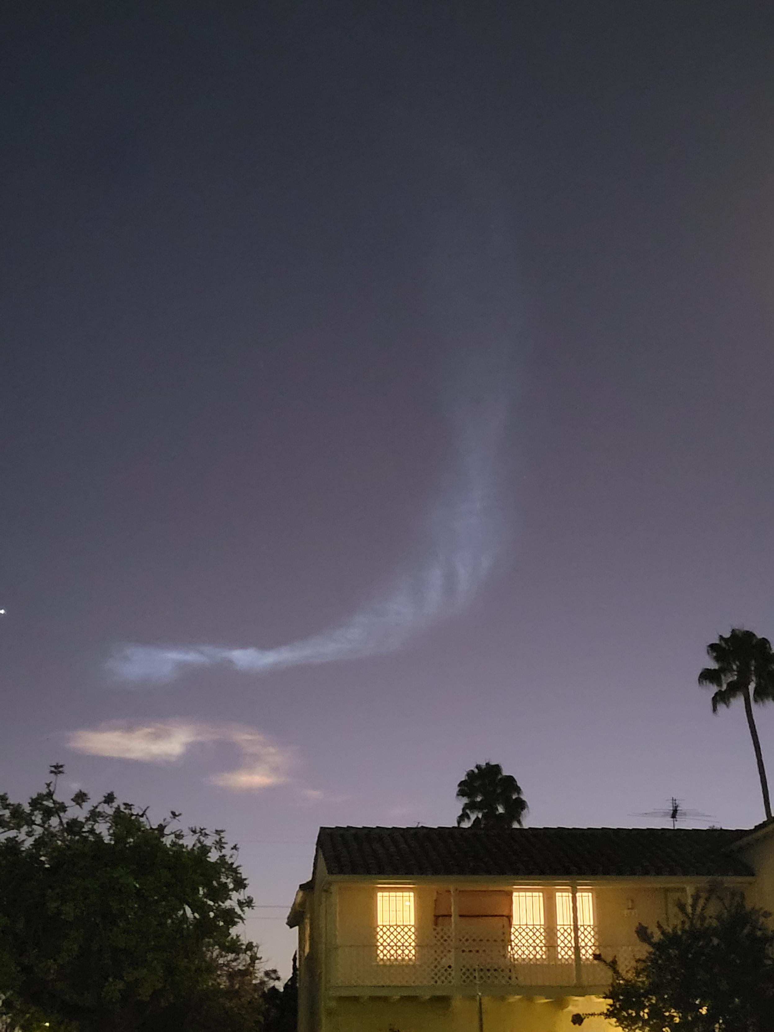 Much better photo of two clouds behind palm trees in an otherwise nearly featureless purple evening sky. The clouds look like wide, swirling doodles, the top one blue and the bottom one pink. To the left of the picture is the lone visible star, which is probably actually the planet Venus. To the right and down, a large house with lights on dominates the street level.