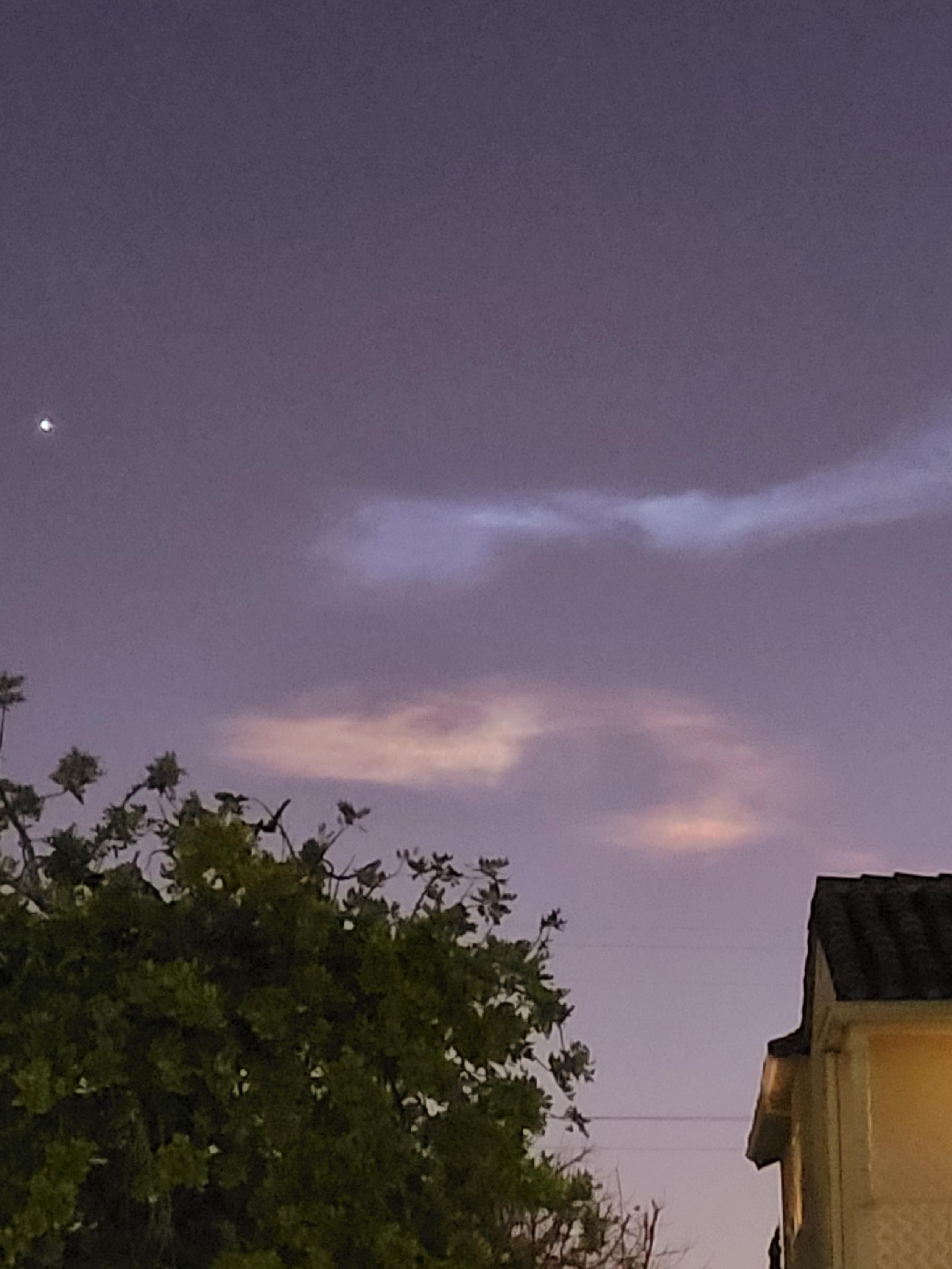 My favorite photo of two noctilucent clouds in an otherwise nearly featureless purple evening sky. The clouds look like wide, swirling doodles, the top one blue and the bottom one pink. The colors are really rich in this one, which captured the scene faithfully. To the left is the lone visible star, which is probably actually the planet Venus. A deciduous tree in full leaf owns the bottom left, with a large pink house filling the bottom right. Wait, that sounds wrong.