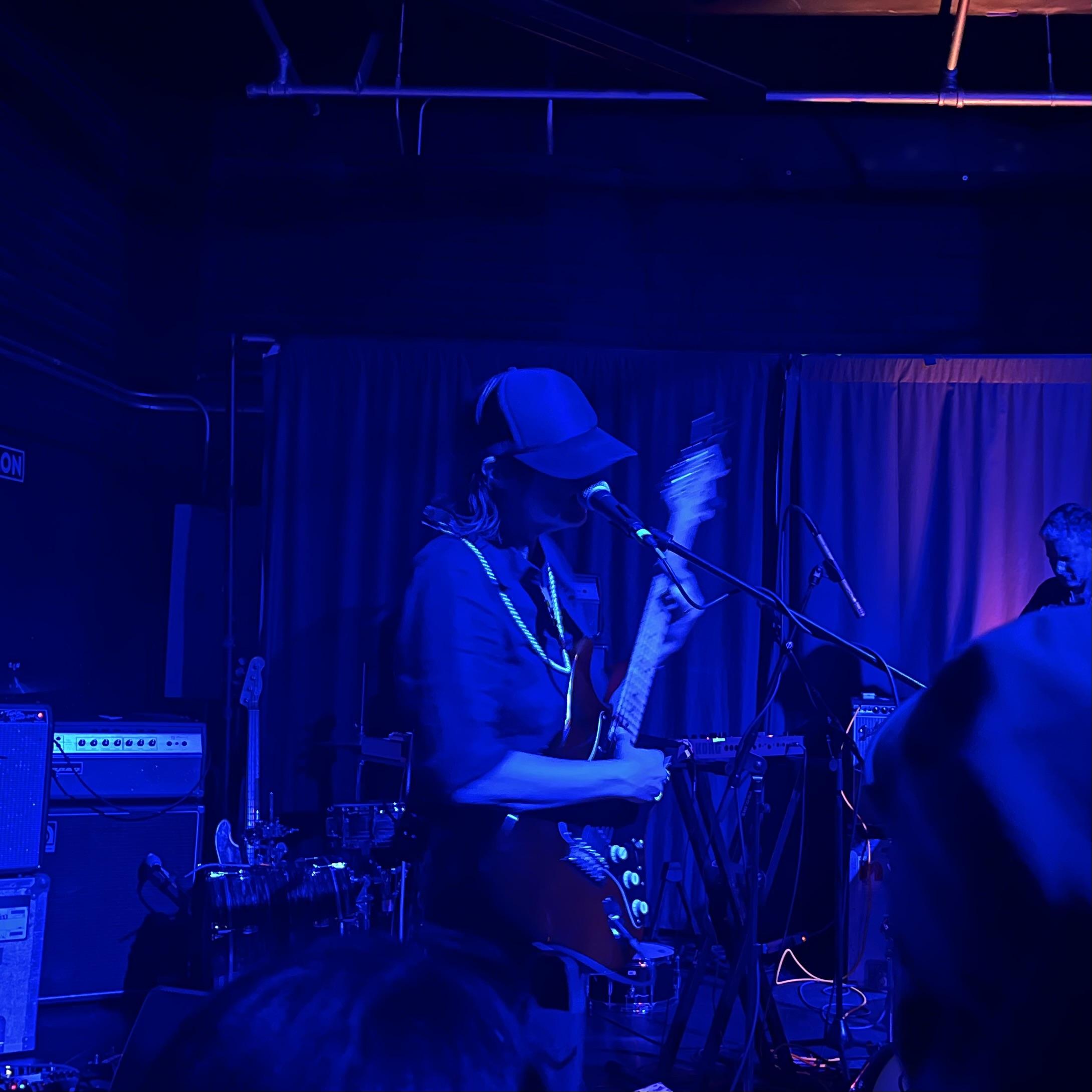 photo of Tara Jane O’Neil playing at Zebulon - dim blue light, wearing a black mesh trucker hat which covers her eyes and playing guitar while singing into the mic