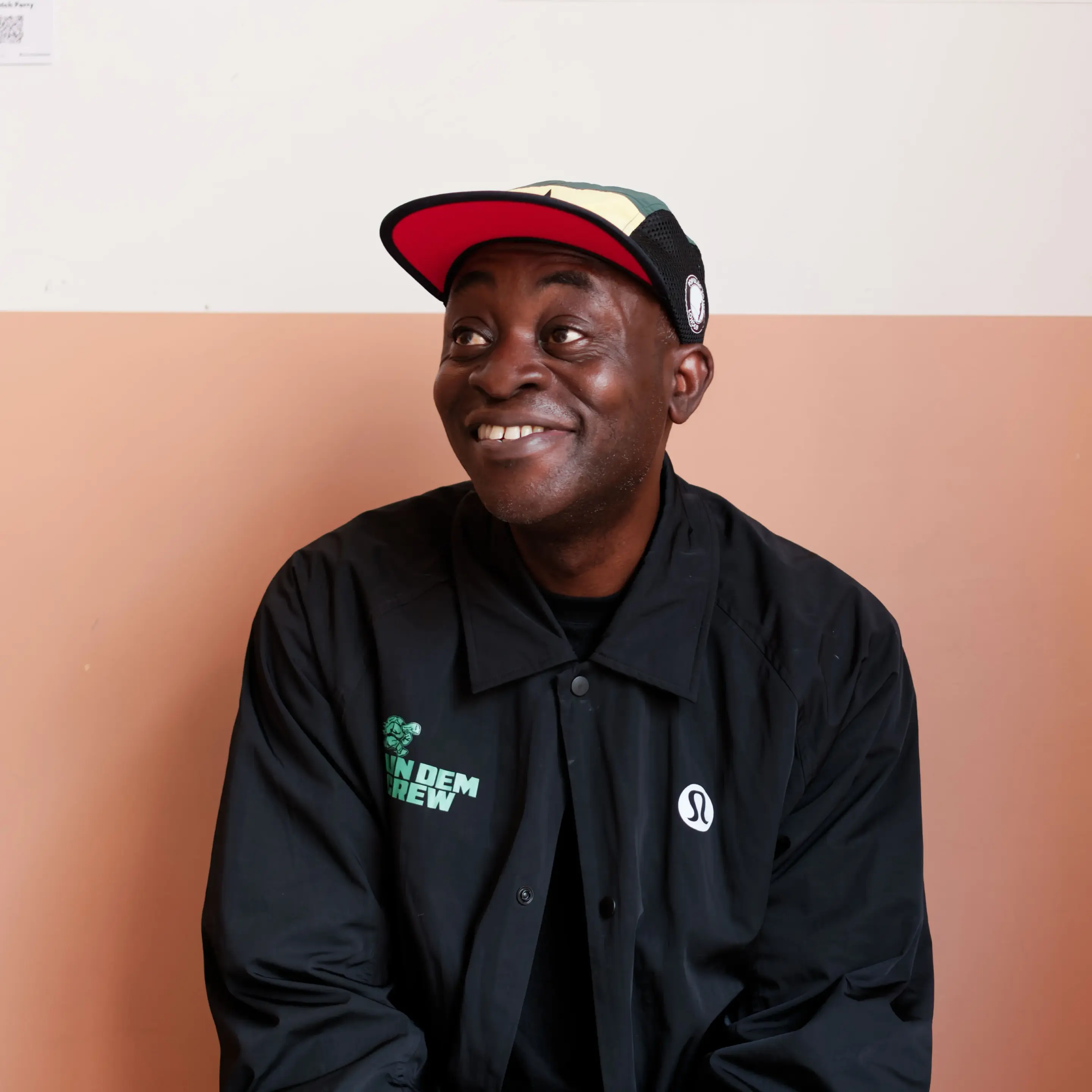 A smiling Black man looking upward and to his left. He wears a black jacket featuring a green logo on the right chest that reads "RUN DEM CREW" with a small turtle icon, and a white Lululemon logo on the left. He is also wearing a cap with a red underbrim, green and yellow top panels, and a round black-and-white patch on the side. He sits in front of a wall split horizontally into off-white and warm peach sections.
