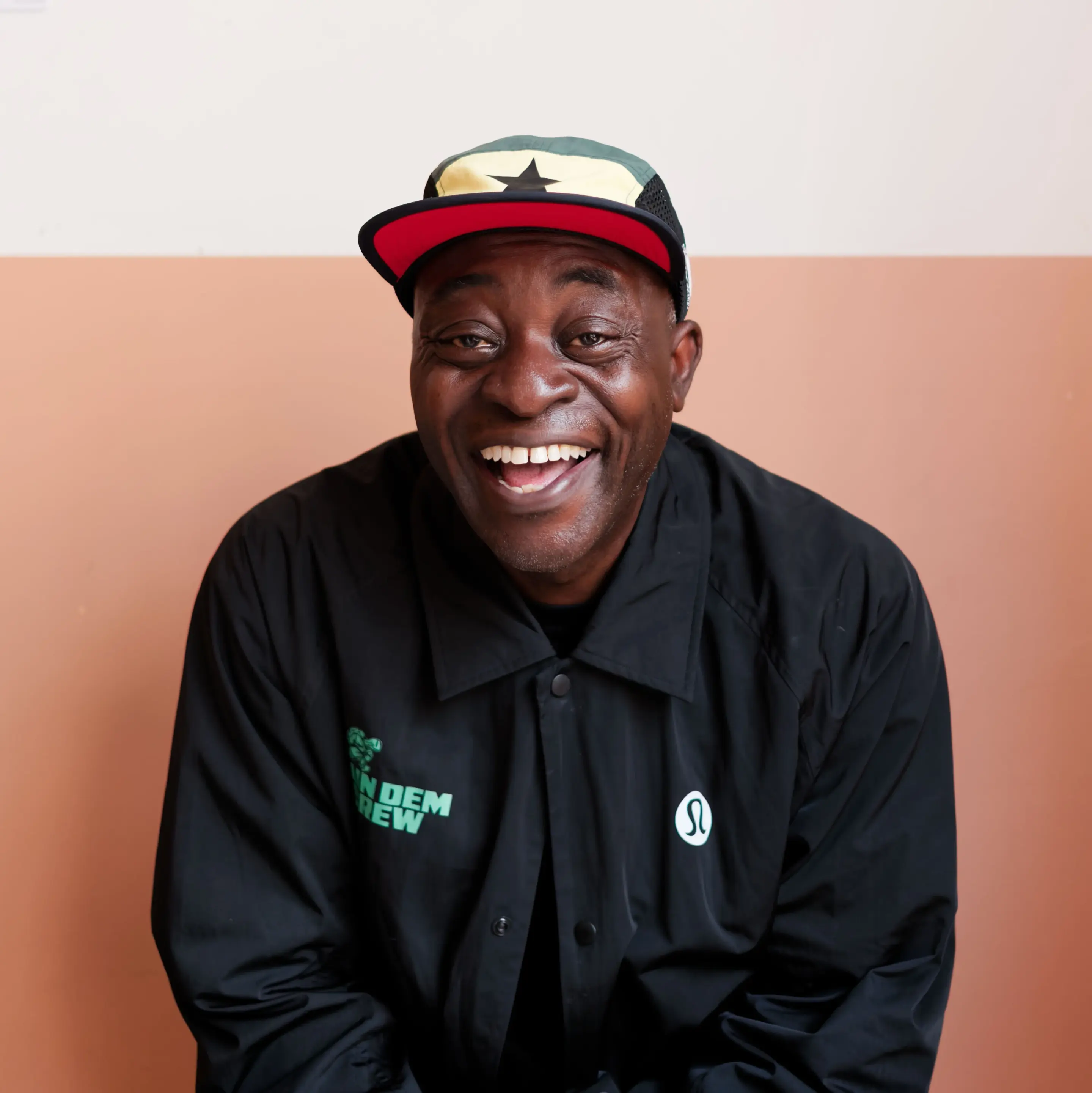 A close-up portrait of a Black man with a joyful, broad smile, looking slightly to his left. He wears a multi-colored cap featuring a green, yellow, and black design on top, a black brim, and a red underbrim. He is dressed in a black button-up jacket that has green text "RUN DEM CREW" on the left chest and a small white stylized "Ω" logo on the right chest. The background is a two-tone wall with a lower section in light terracotta and an upper section in off-white.