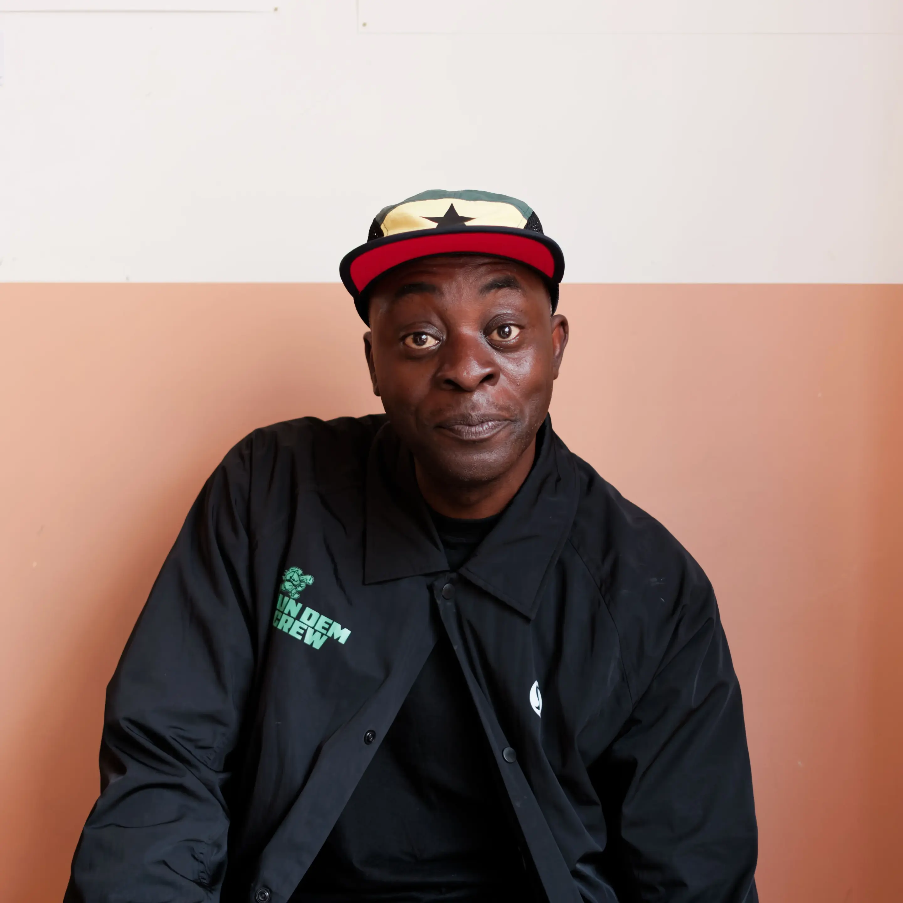 A Black man with a gentle expression looking directly at the camera with wide eyes and closed lips, tilted slightly to his right. He wears a black button-down jacket over a dark shirt, featuring green text that reads "RUN DEM CREW" with a turtle icon on the right side and a white circular logo on the left. His cap has a red underbrim, a green and yellow crown panel with a black star in the center, and black mesh side panels. The background is a wall split horizontally into an off-white top section and a warm peach bottom section.