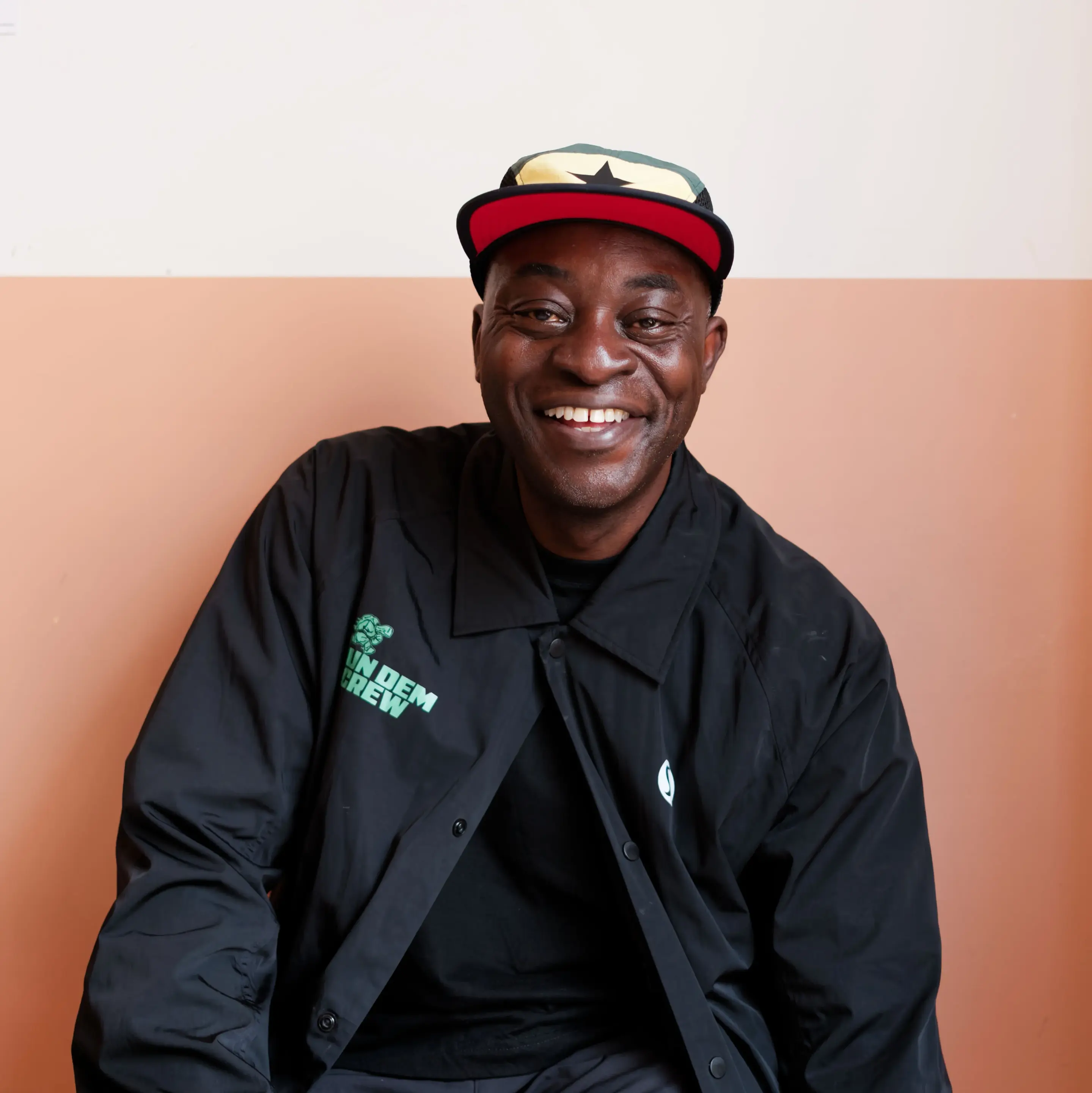 A Black man with a broad, warm smile facing the camera. He is wearing a black button-up jacket over a dark top, with a green logo reading "RUN DEM CREW" alongside a small turtle graphic on the right chest and a white circular logo on the left. His cap features a yellow front panel with a black five-pointed star, green top, red underbrim, and black mesh side panels. He sits centered against a two-tone wall with off-white above and warm terracotta below.